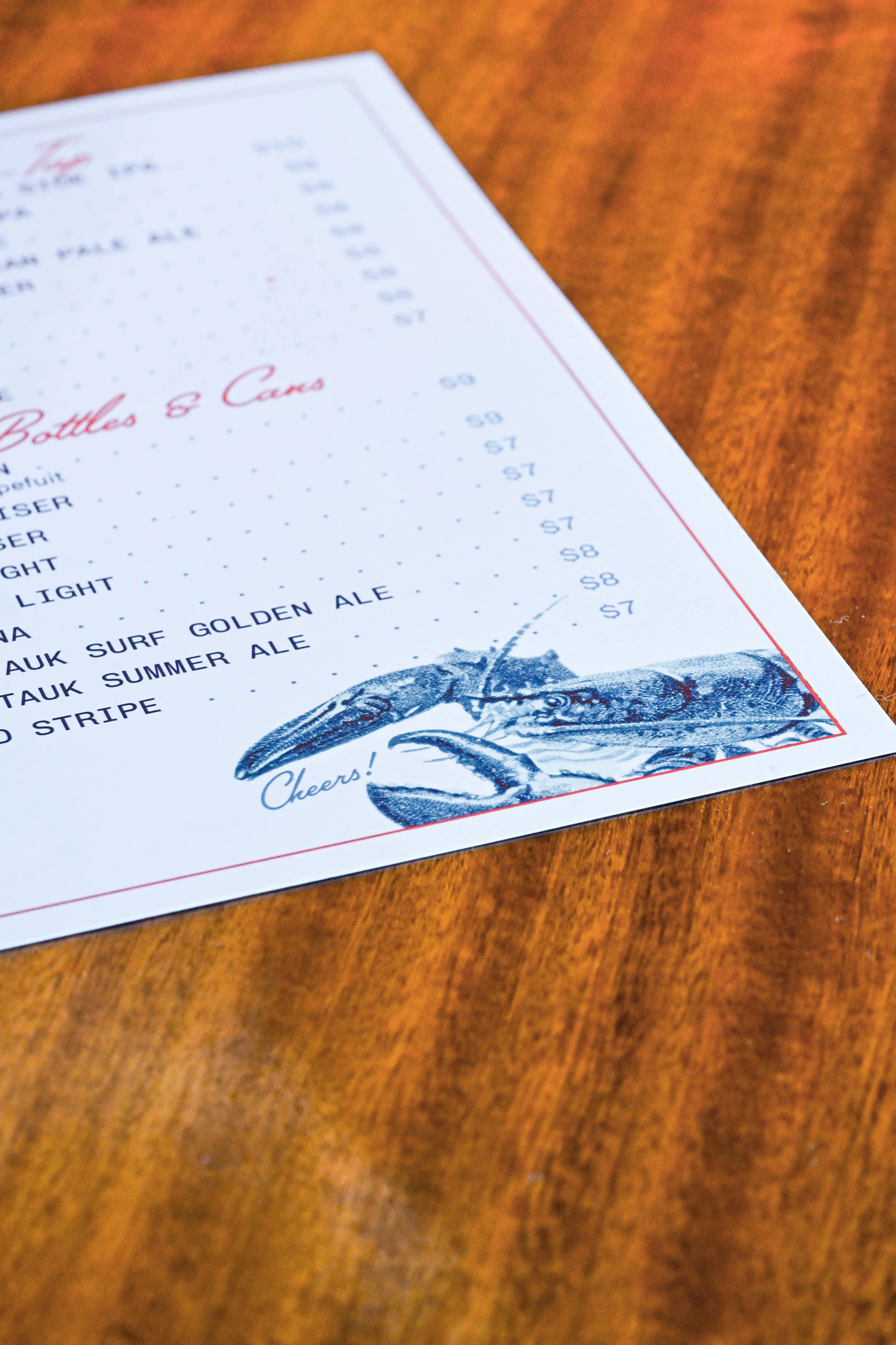 Close-up of a menu on a wooden table featuring a drawing of a lobster saying "Cheers" at the bottom.