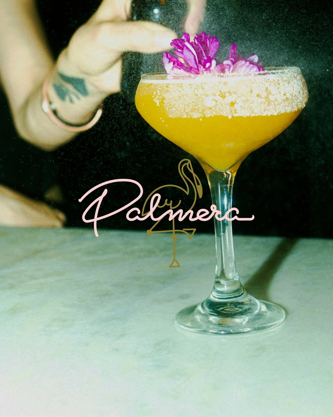 A yellow cocktail in a coupe glass with a salted rim, garnished with purple flowers with the 'Palmera' logo across the image.