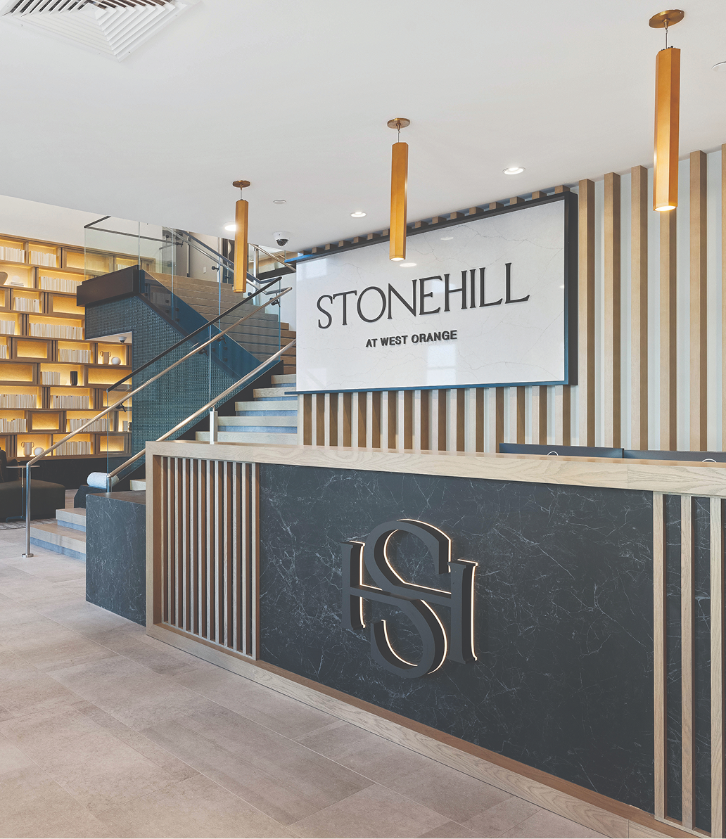 Modern apartment lobby with a front desk featuring a black marble facade and a silver 'S' logo, wooden slats decor, a large sign that reads 'STONEHILL AT WEST ORANGE,' and a staircase with wooden treads and glass railings leading to the upper level.