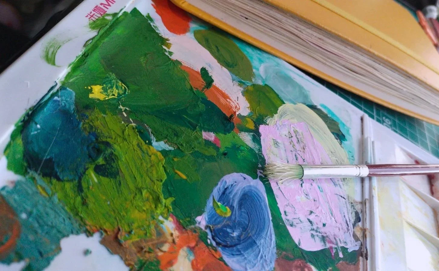 Paleta con pintura seca, el pincel y el sketchbook o cuaderno de dibujos al lado. Hay diferentes colores que muestran una paleta variada y vibrante.