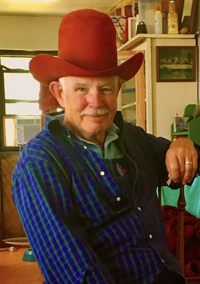 An older man wearing a brown cowboy hat, blue plaid shirt, and dark vest, smiling indoors with various kitchen items in the background.