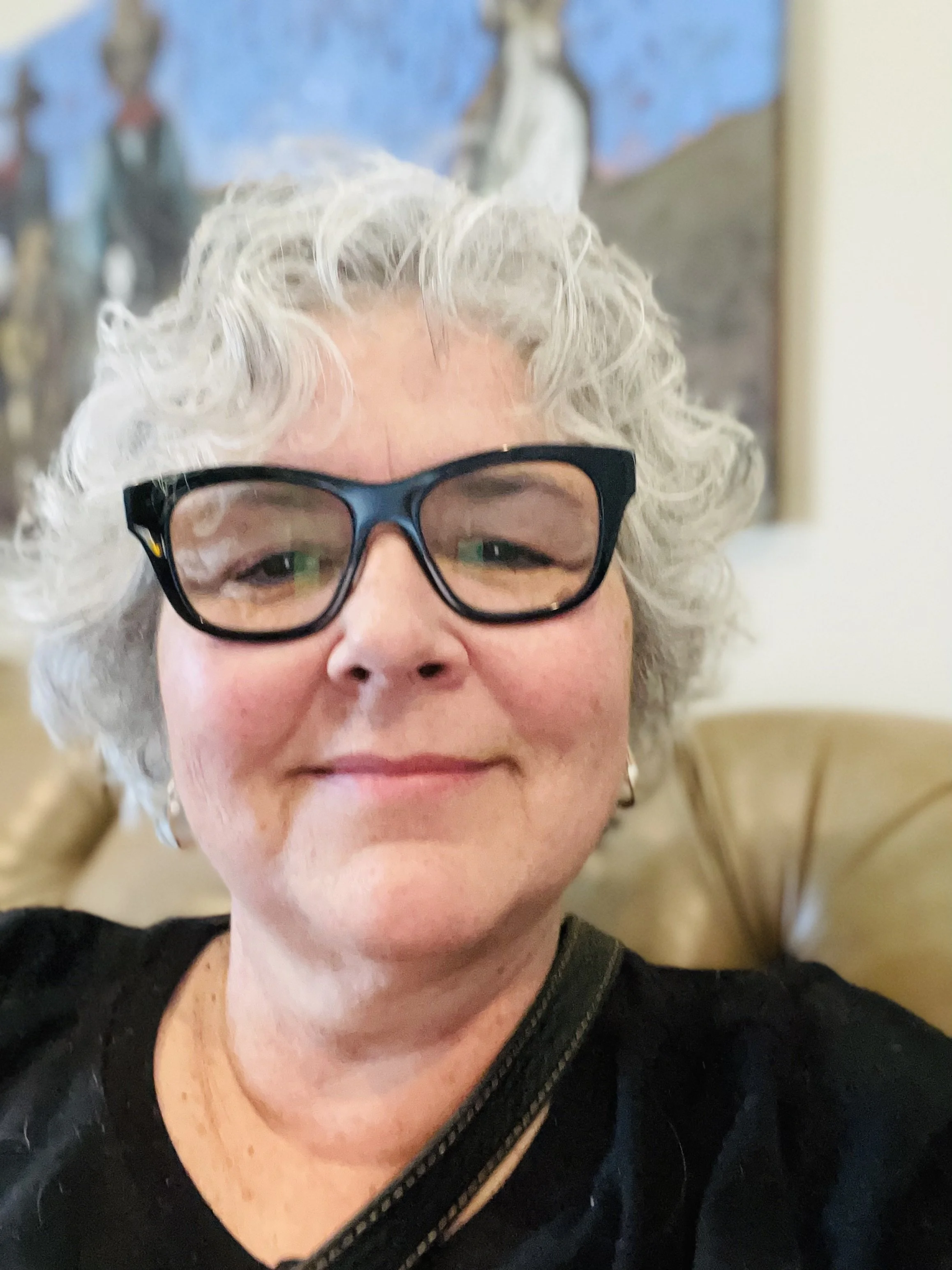 A woman with curly gray hair, wearing glasses, smiling and sitting in a room with a blue-colored background.