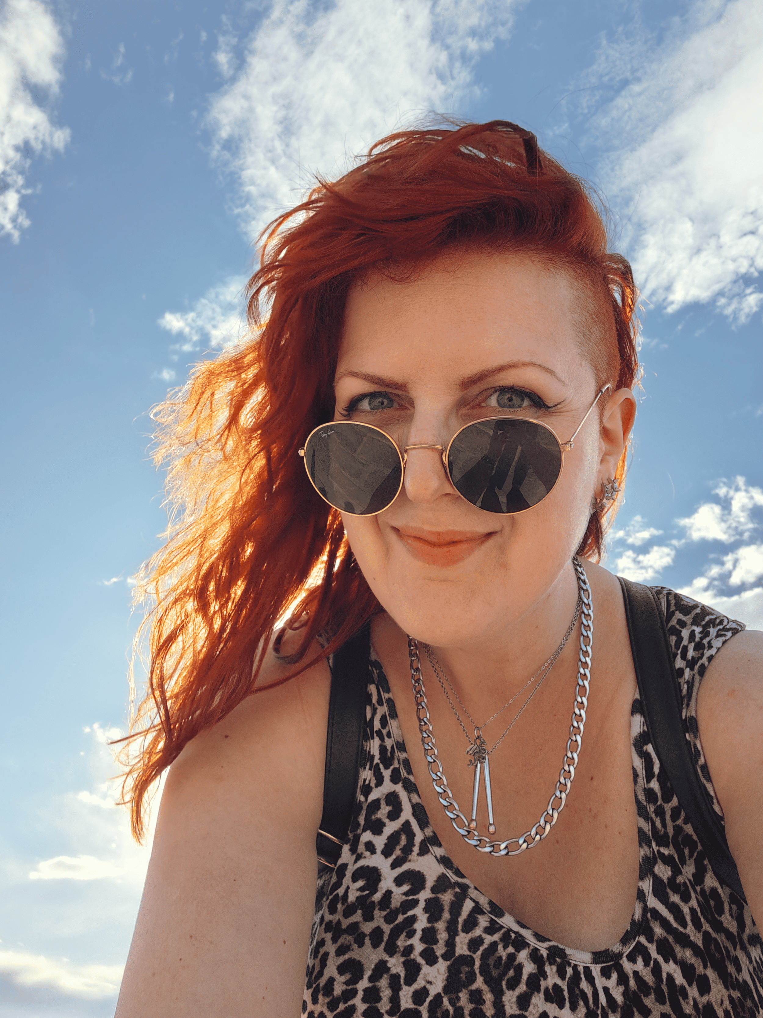 A woman with red hair (Scilla, pronounced Shilla) wearing round sunglasses, a leopard print top, silver chains, and earrings, smiling outdoors with a blue sky and clouds in the background.