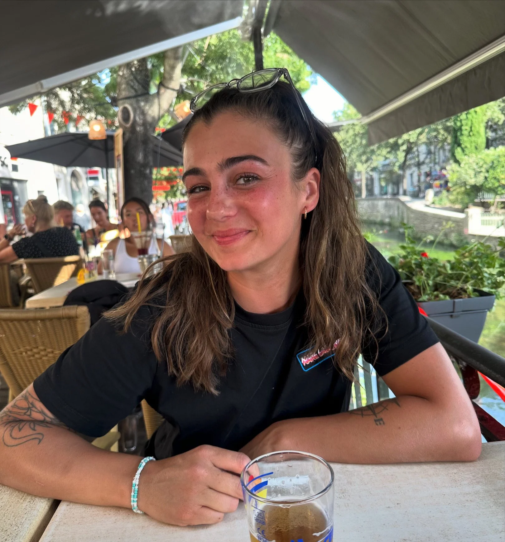 Jeune femme assise à une table d'un restaurant en plein air, souriant, avec une boisson devant elle, portant des lunettes sur la tête, dans une ambiance estivale.