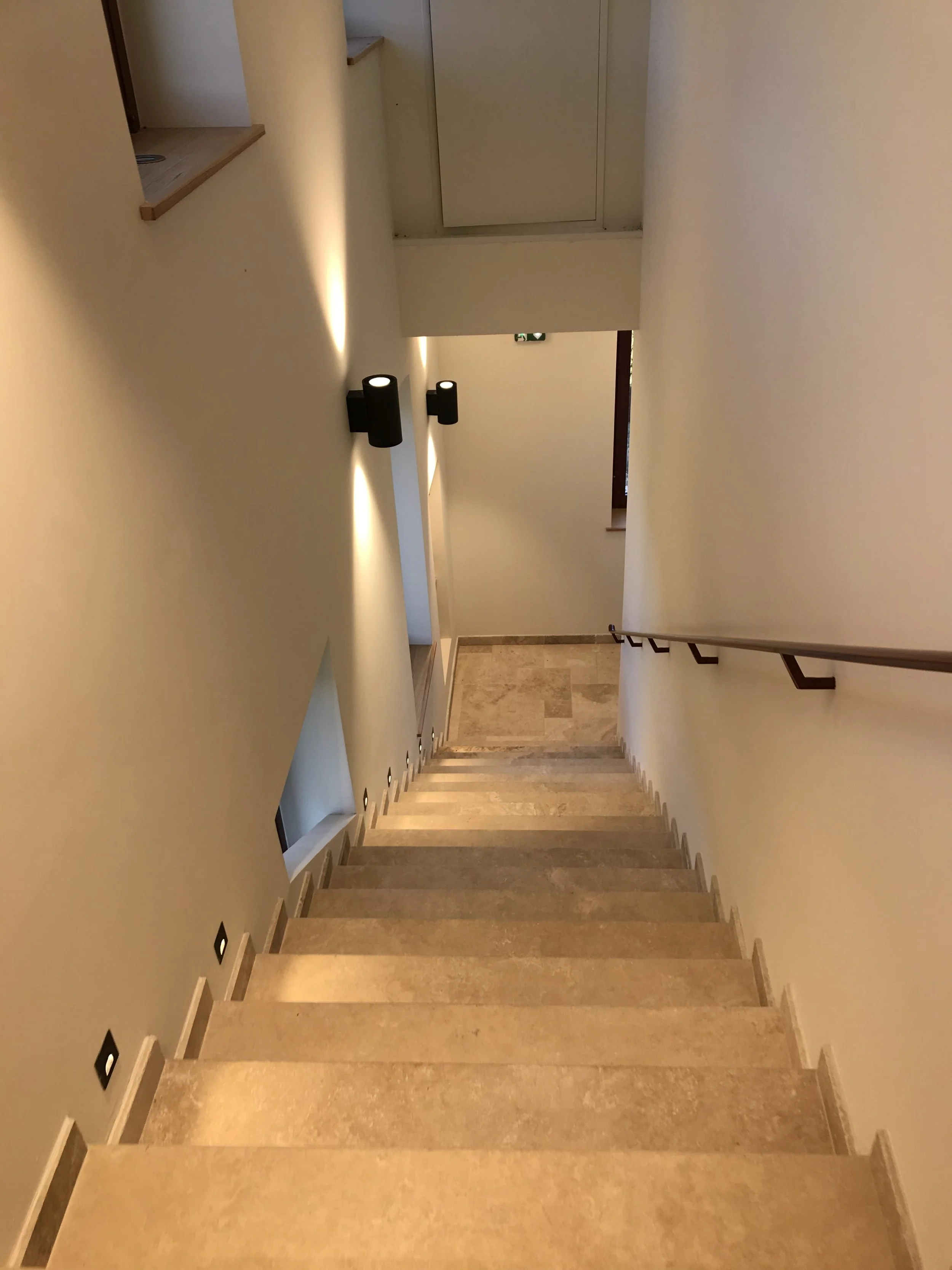 Photo d'un escalier en pierre beige avec mur blanc, éclairé par des lampes murales noires.