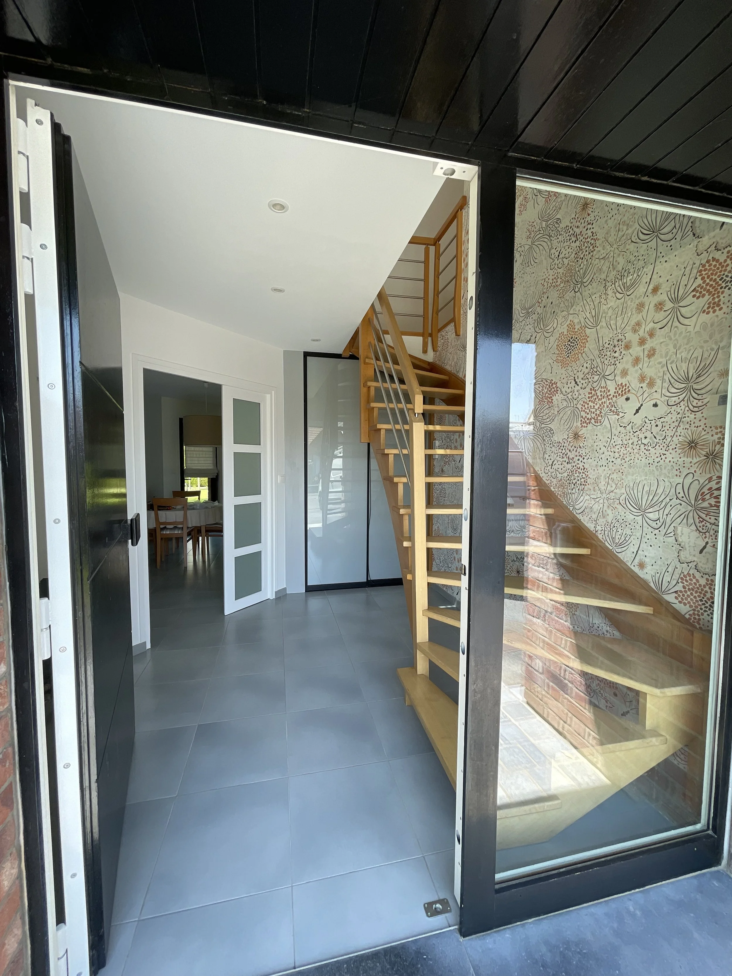 Intérieur d'une maison avec un escalier en bois, un mur avec un papier peint floral, une porte vitrée, et une salle à manger visible au fond.