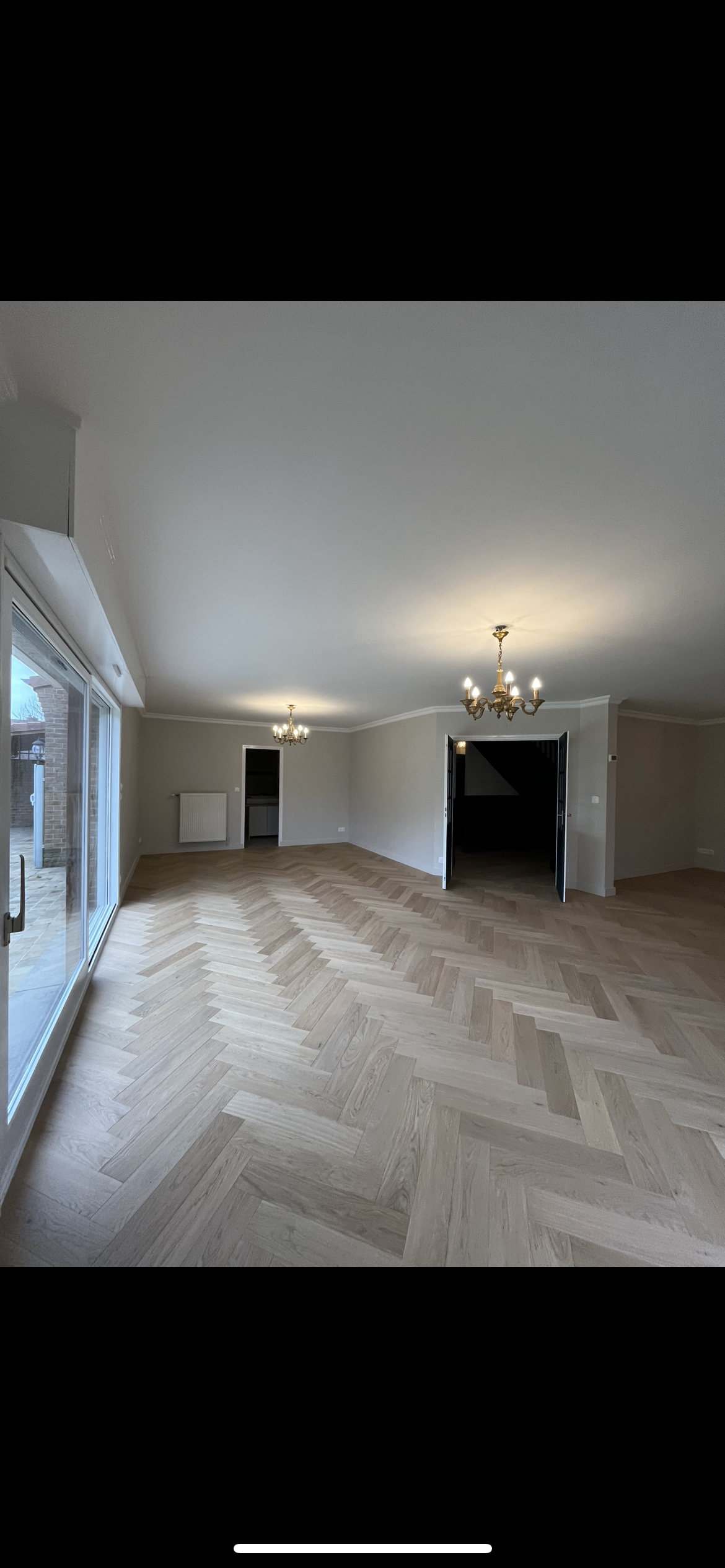 Une pièce vide avec un parquet en bois clair en motif chevron, des lustres suspendus, une grande porte-fenêtre en verre avec volet roulant, et des murs peints en blanc ou beige. Il y a une ouverture menant à une autre pièce.