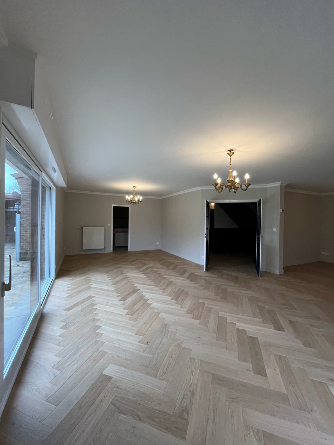 Grand salon avec parquet en bois clair, deux lustres en or, porte-fenêtre donnant sur l'extérieur, murs blancs, espace vide.