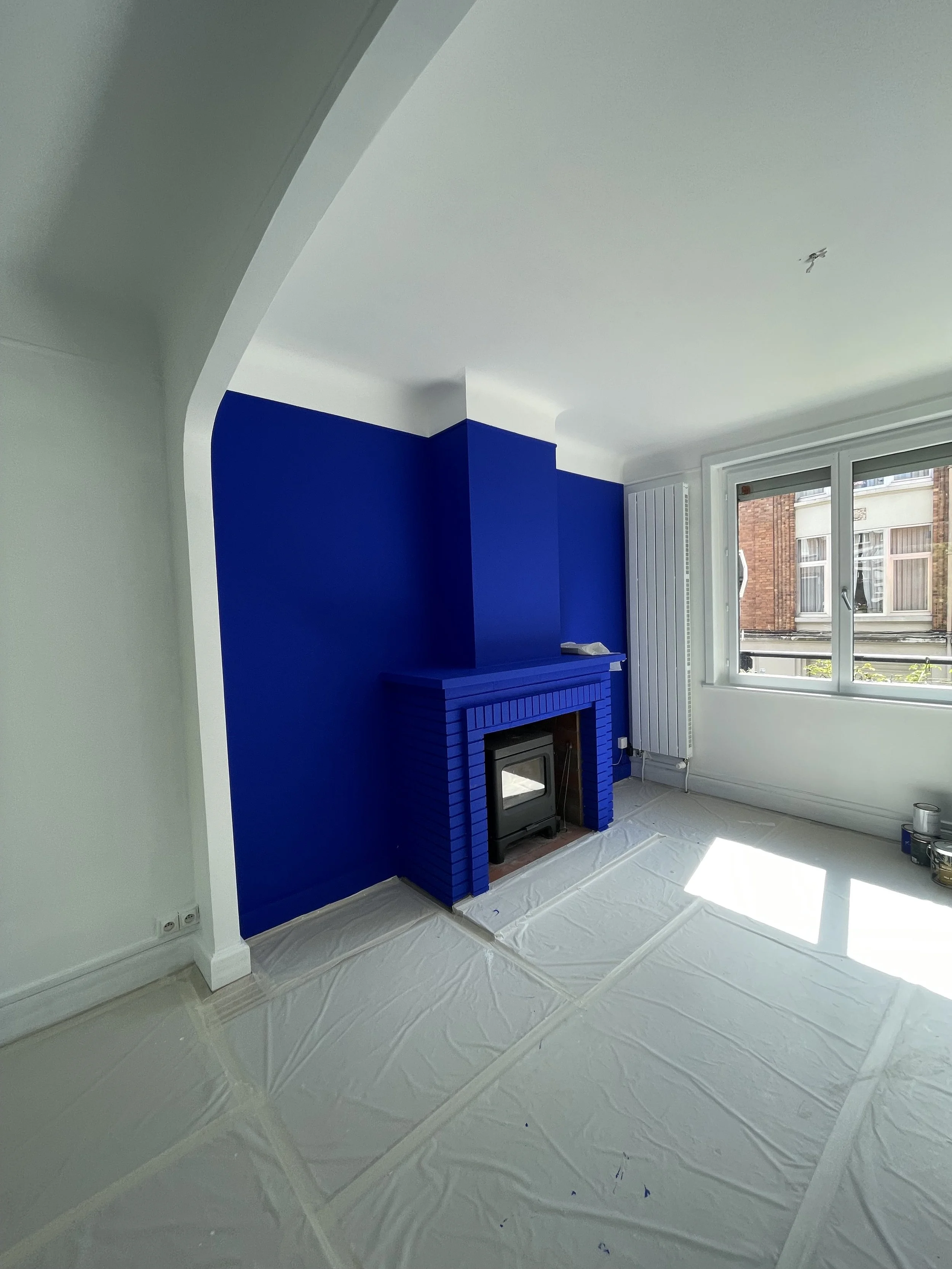 Intérieur d'un salon en cours de décoration avec un mur peint en bleu, un foyer, une grande fenêtre laissant entrer la lumière naturelle, et des surfaces protégées par des draps.