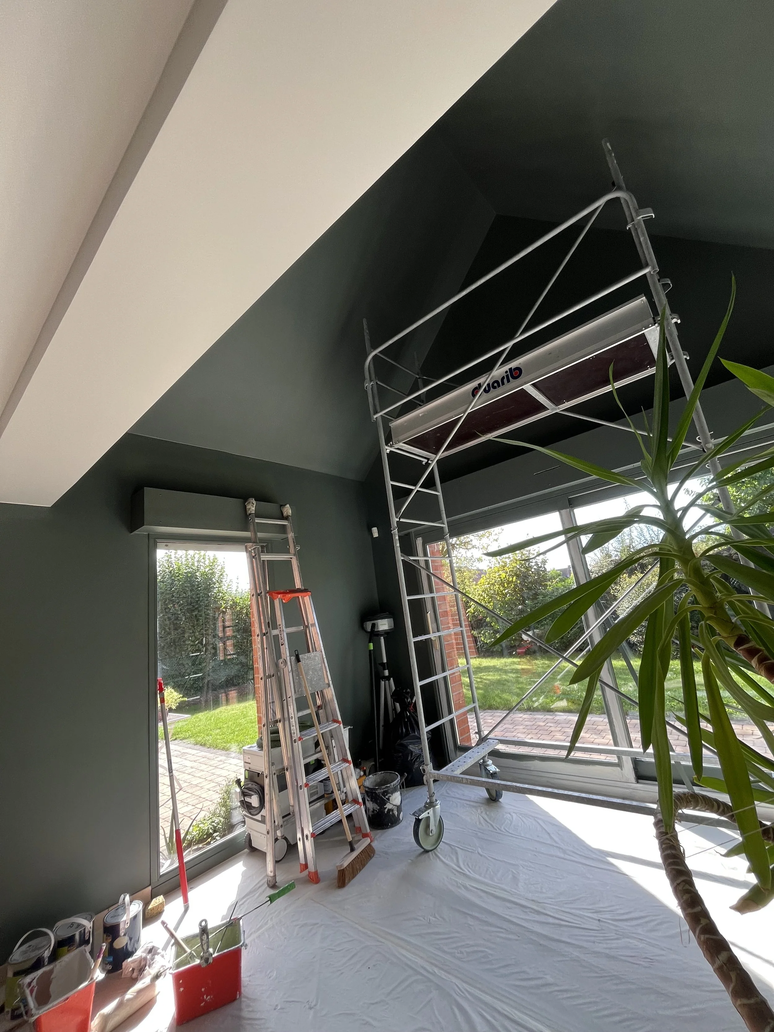 Intérieur d'une pièce en cours de rénovation avec un escalier roulant et une échelle, près d'une grande fenêtre donnant sur un jardin ensoleillé, avec divers outils et matériaux de peinture et de bricolage.