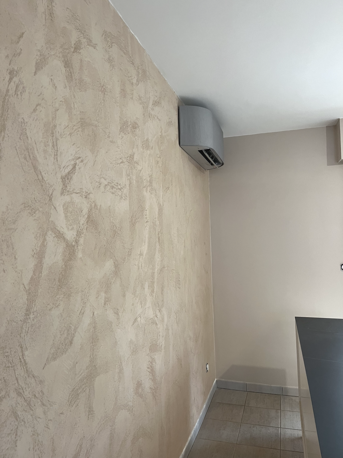 Mur beige avec texture rustique, ventila­tion encastrée au plafond, sol en carreaux, coin de mur avec comptoir noir en stratifié.