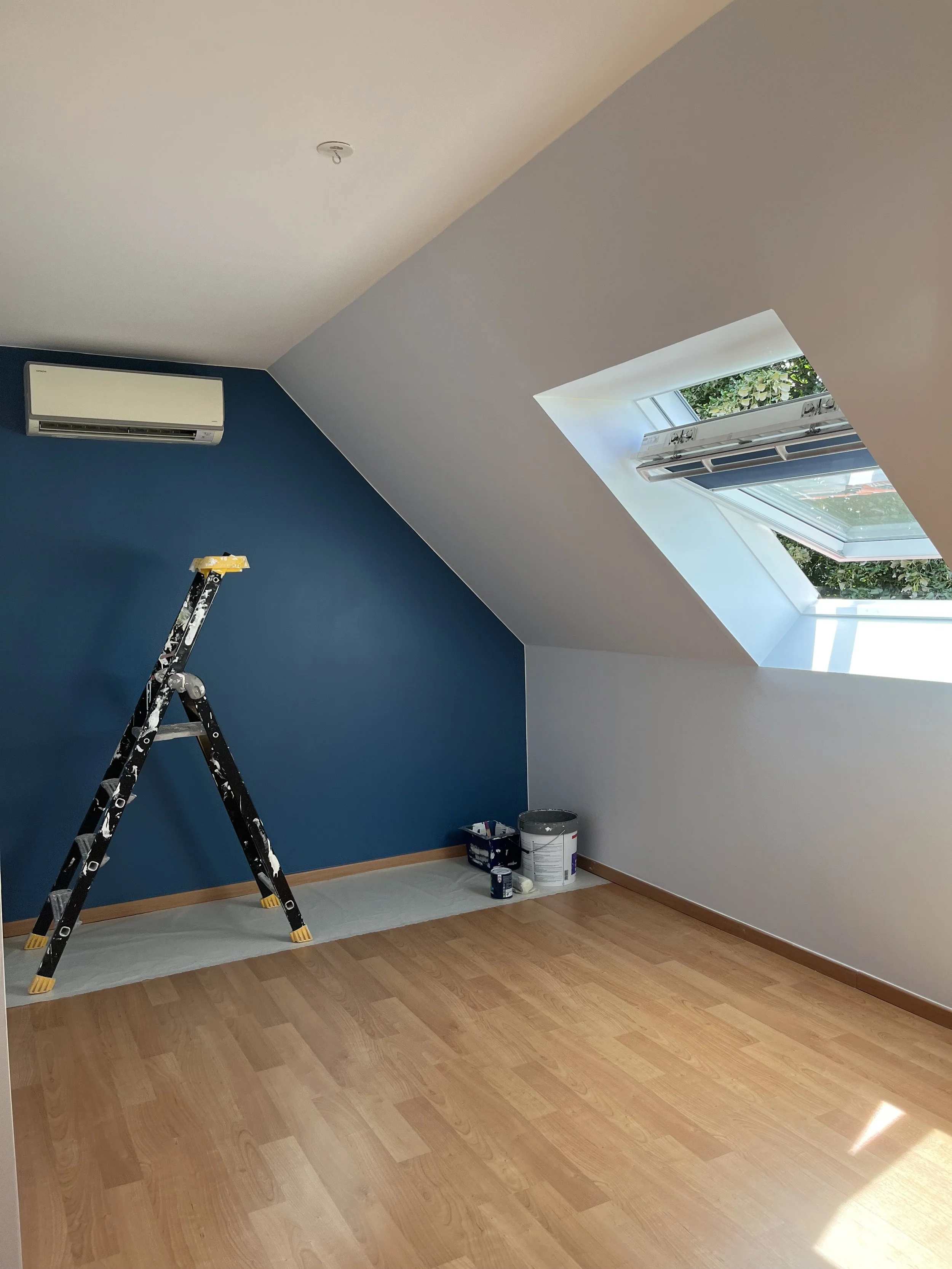 Une pièce en cours de peinture avec un mur bleu, un plafond blanc avec une fenêtre de toit ouverte, une échelle noire, et des pot de peinture dans un coin.