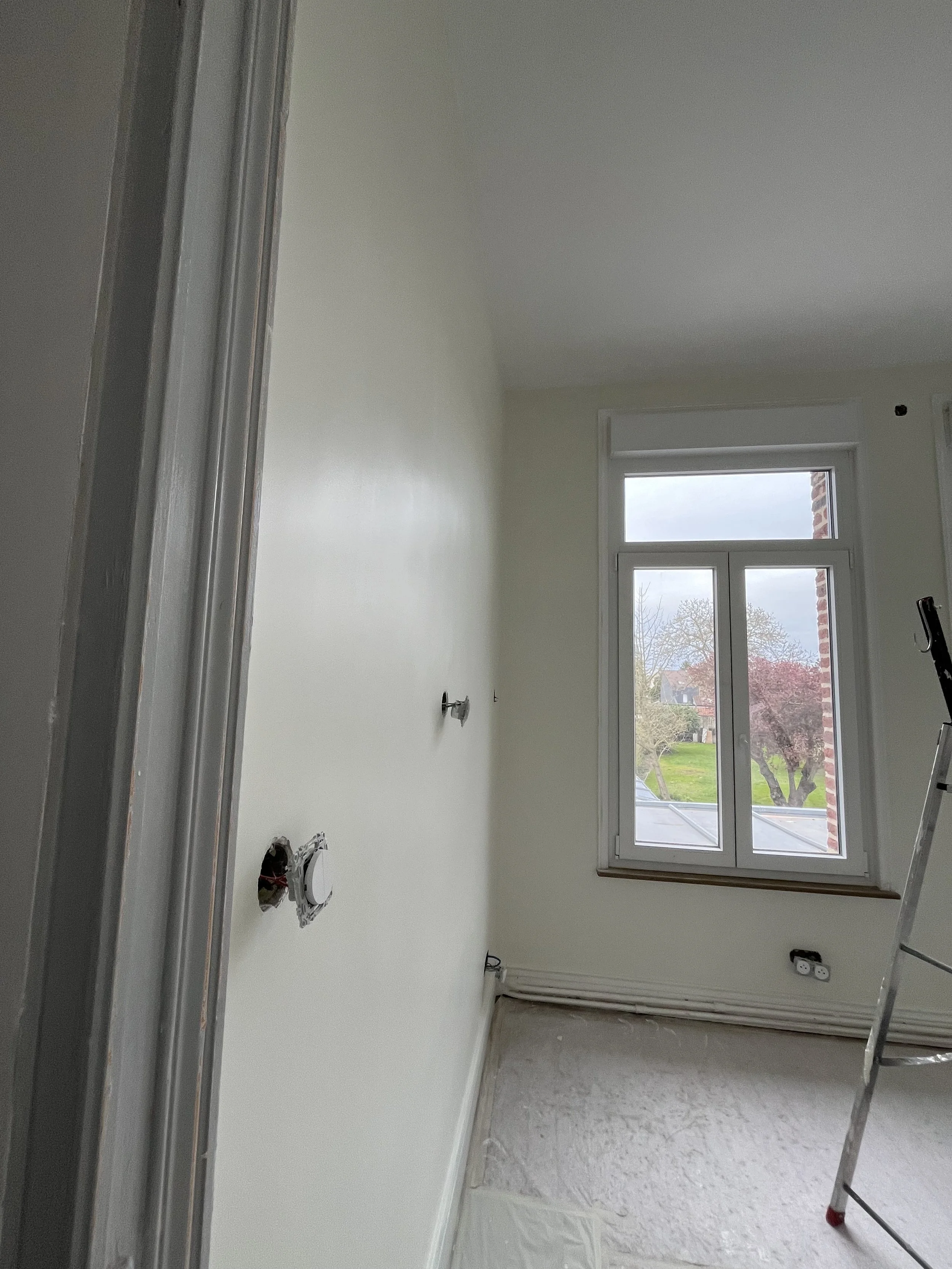 Une pièce en cours de rénovation avec un mur peint en blanc, une fenêtre donnant sur un jardin, un échelle en métal et des câbles électriques au sol.