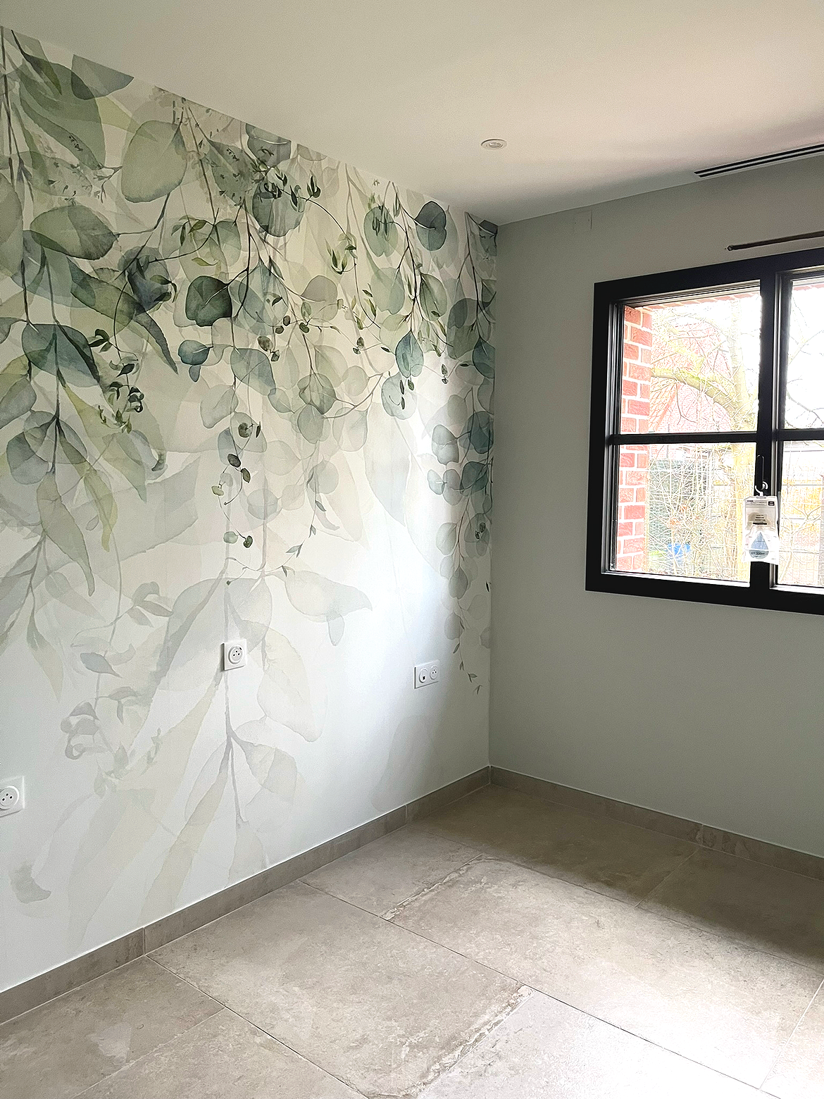 Une pièce avec un mur décoré d'un papier peint à motifs de feuilles et de branches, une fenêtre avec cadre noir laissant entrer la lumière naturelle.