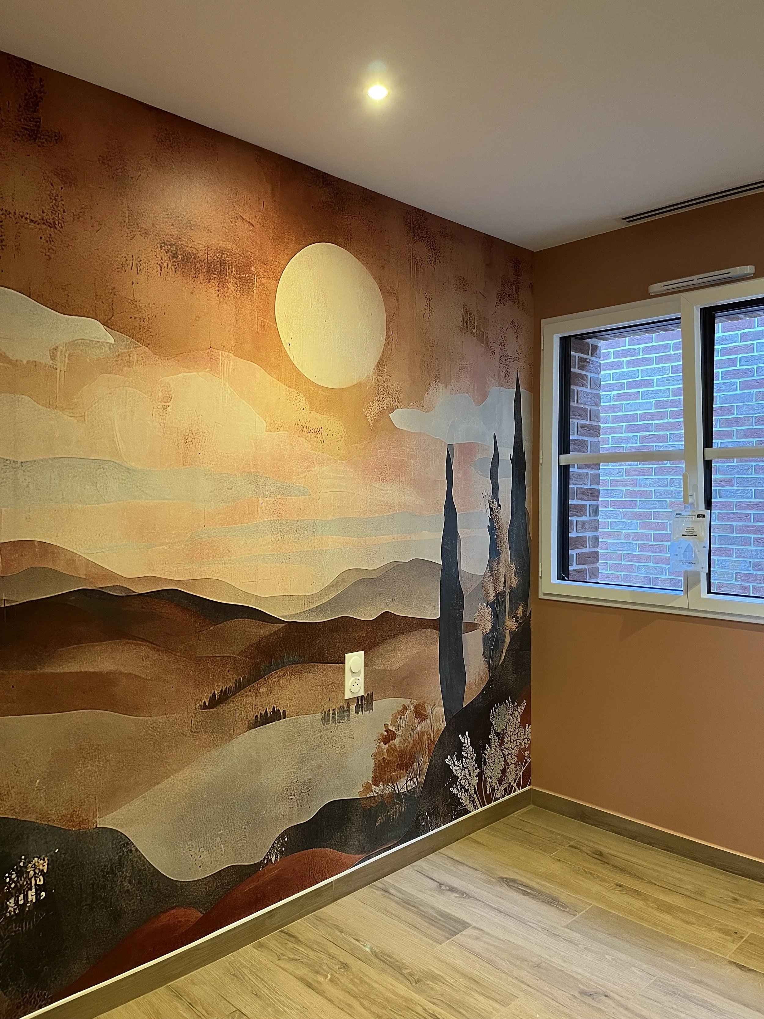 Mur peint avec une fresque représentant un paysage de montagne avec un soleil, des arbres, et un ciel aux couleurs chaudes, dans une pièce avec un sol en bois et une fenêtre.