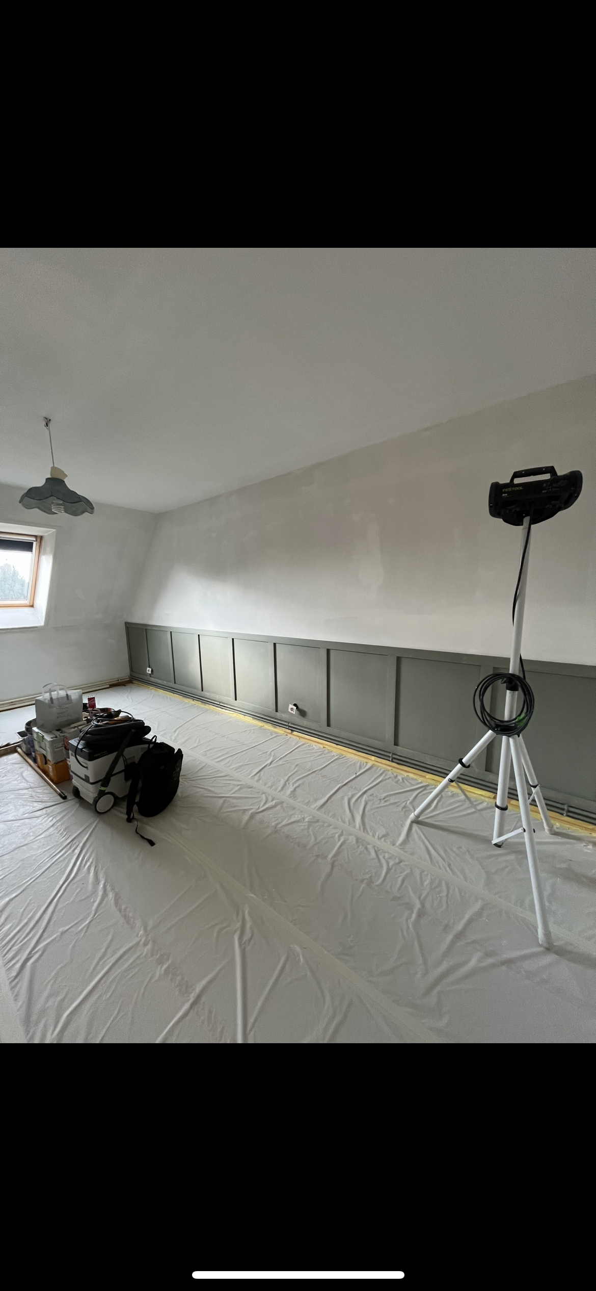 Pièce en cours de rénovation avec un mur récemment peint, une lampe suspendue au plafond, une fenêtre, un espace recouvert de plastique pour la peinture, et des équipements de travail.