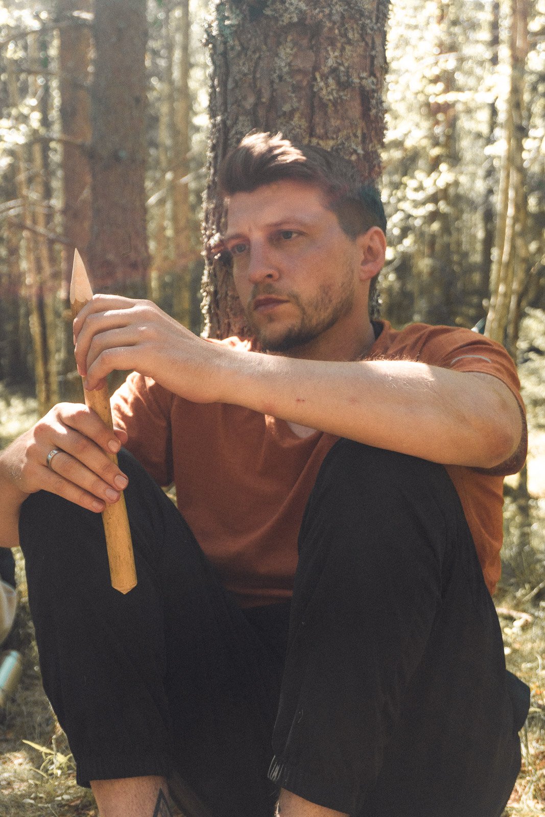 A man with dark hair and a beard sitting in a forest, holding a small axe, with trees and sunlight in the background.
