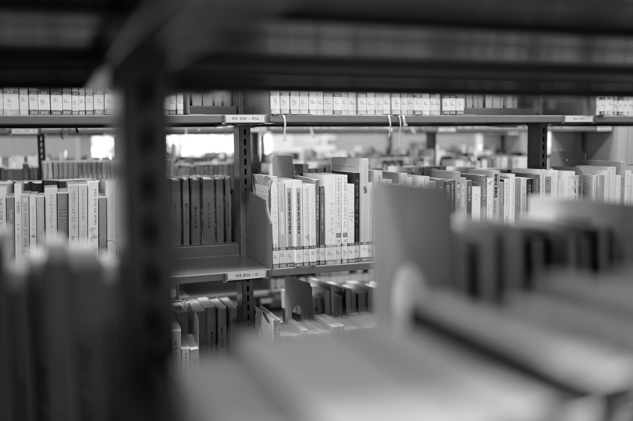 Black and white photo of bookshelves filled with books in a library, viewed through a gap between shelves.