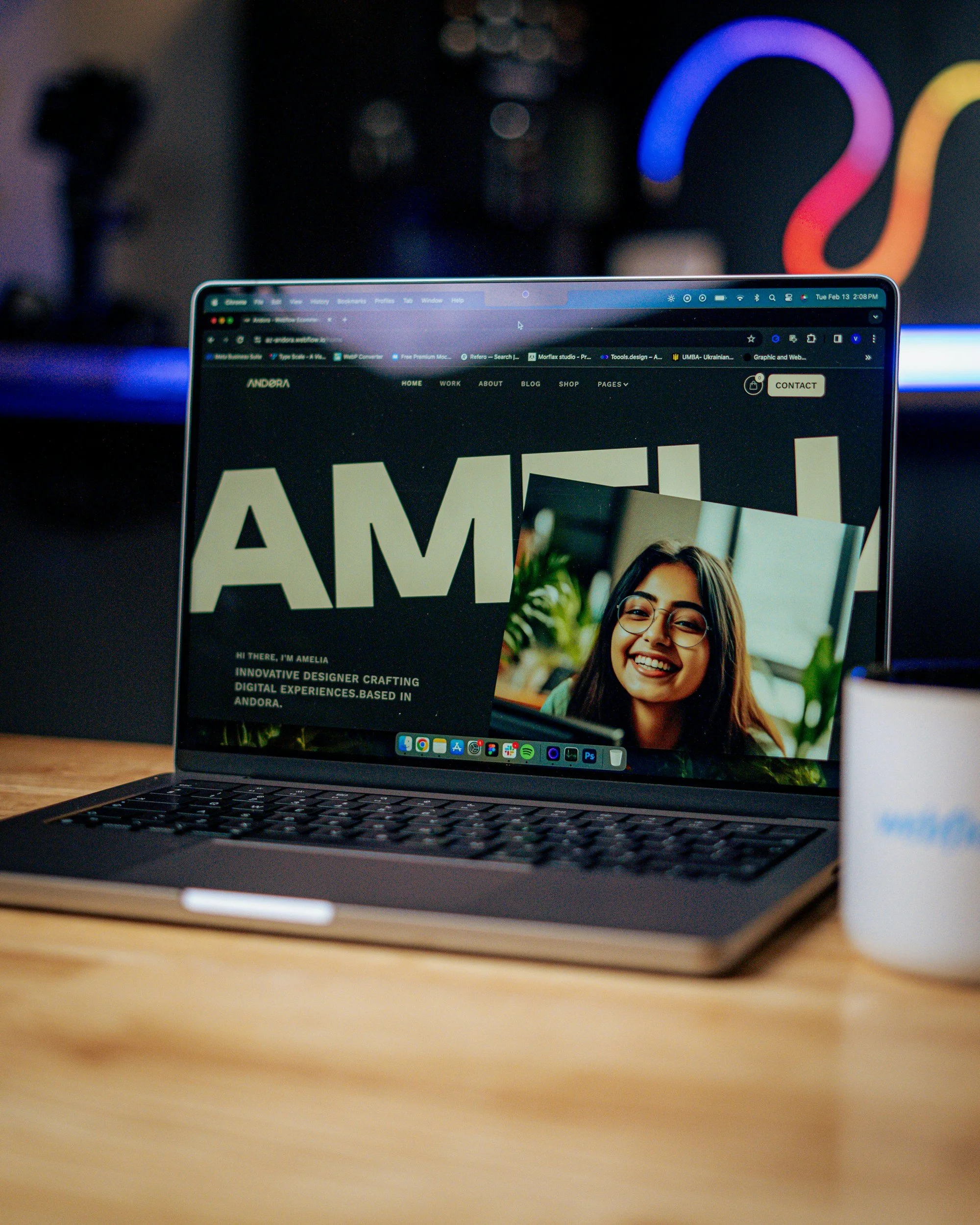 A close-up view of a laptop computer on a wooden desk, displaying a website with the name 'ANDORA' and a photo of a smiling woman. The background is blurred with a dark and colorful light display visible.