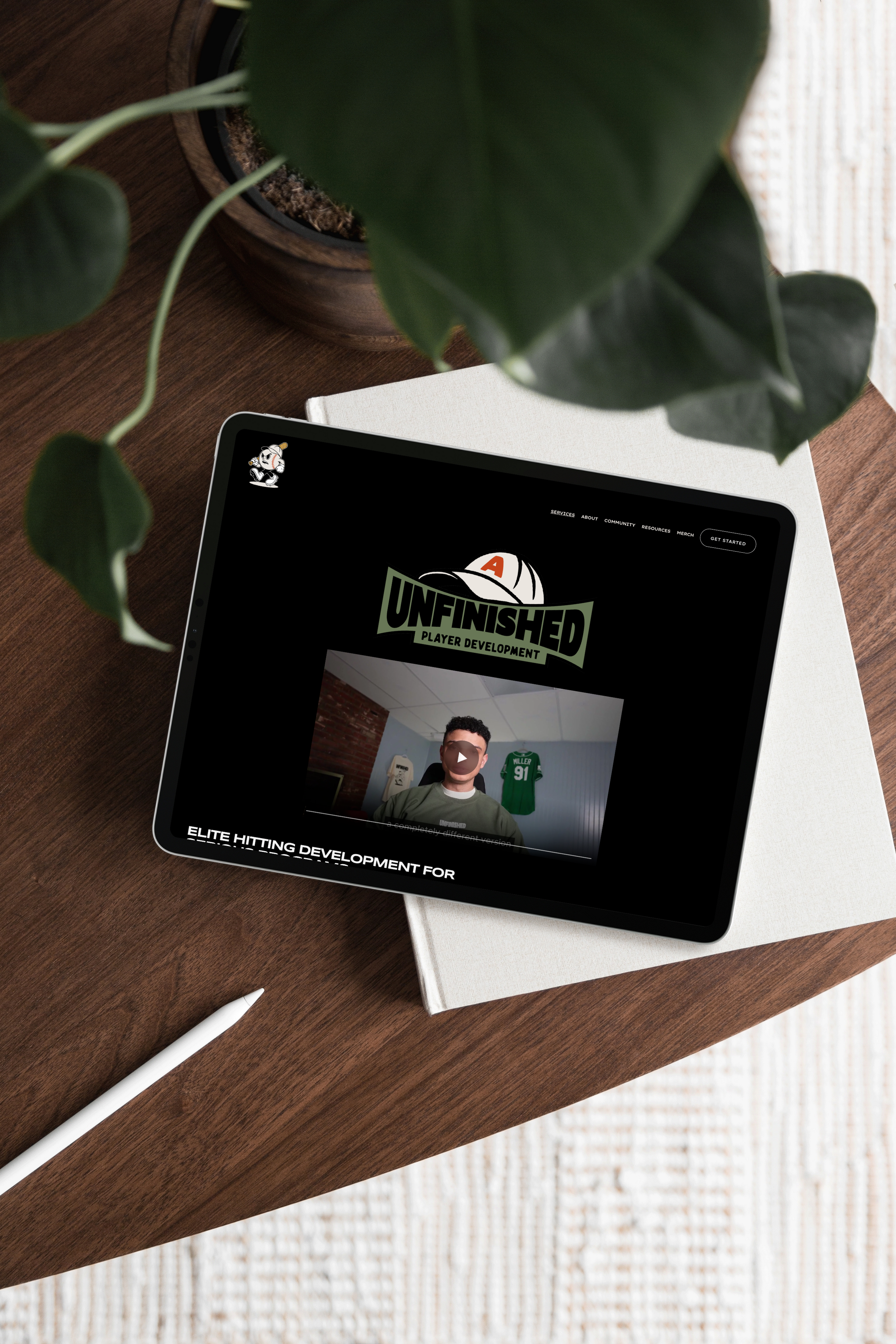 Tablet on a wooden table displaying a sports-themed website with the text "Unfinished Player Development" and an image of a person in front of sports memorabilia, partially covered by a large green leafy plant.