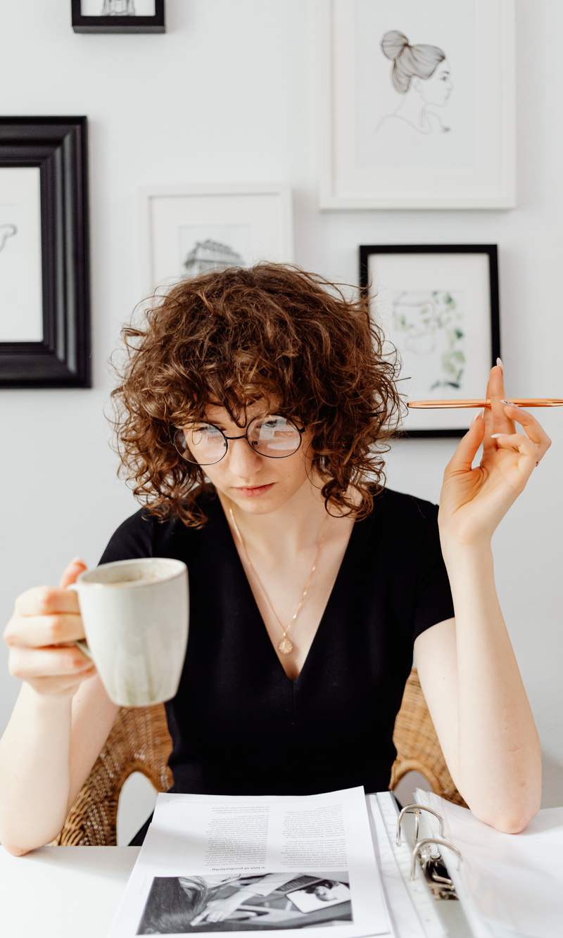 Woman with curly hair and glasses sitting at a desk with papers, holding a mug in one hand and a pen in the other, in a room with framed art on the wall.