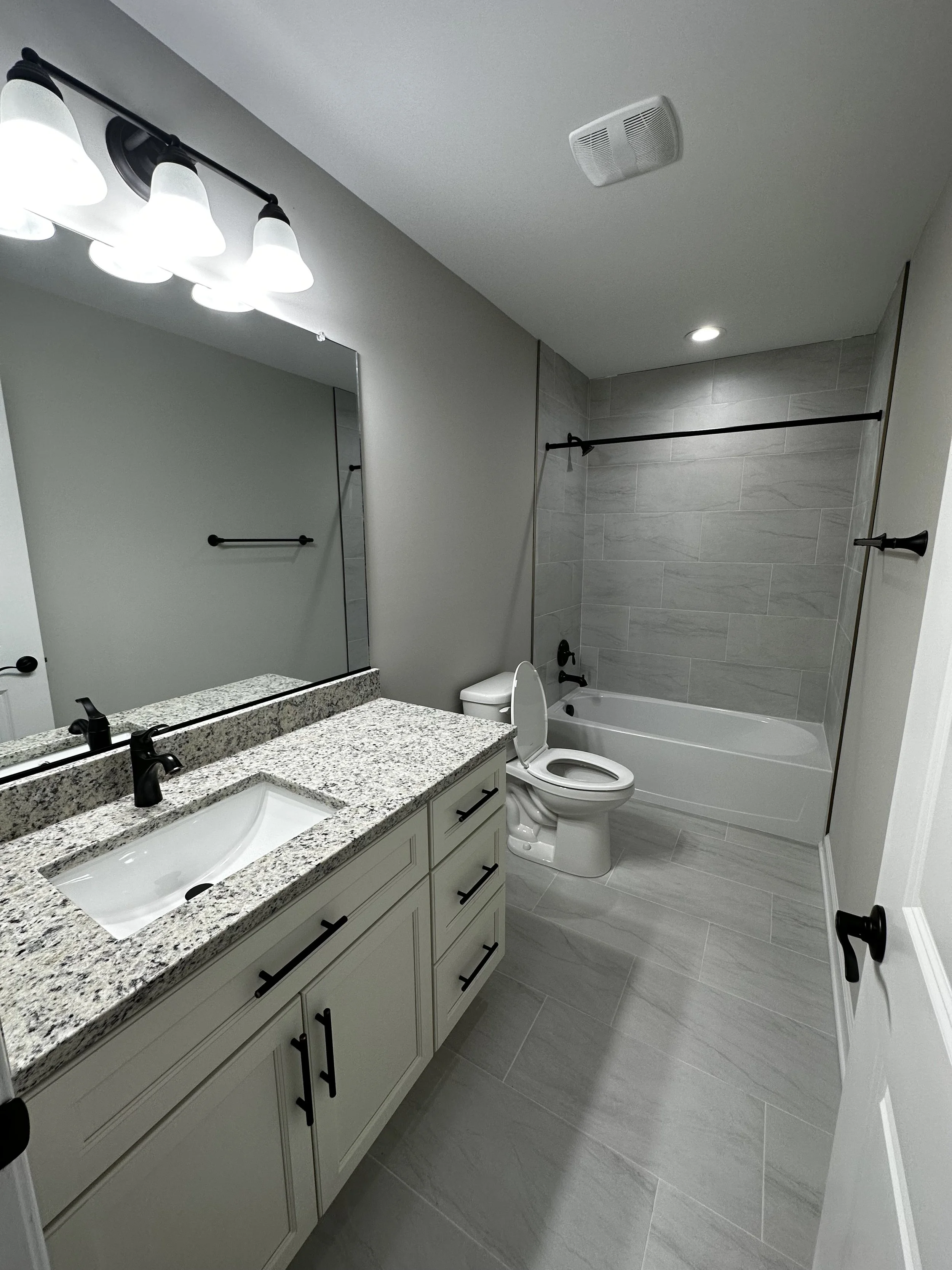 Modern bathroom with light gray tiled flooring and walls, a white vanity with a granite countertop, black faucet and hardware, a large mirror, a toilet, and a bathtub with a shower, black curtain rod, and gray wall tiles.
