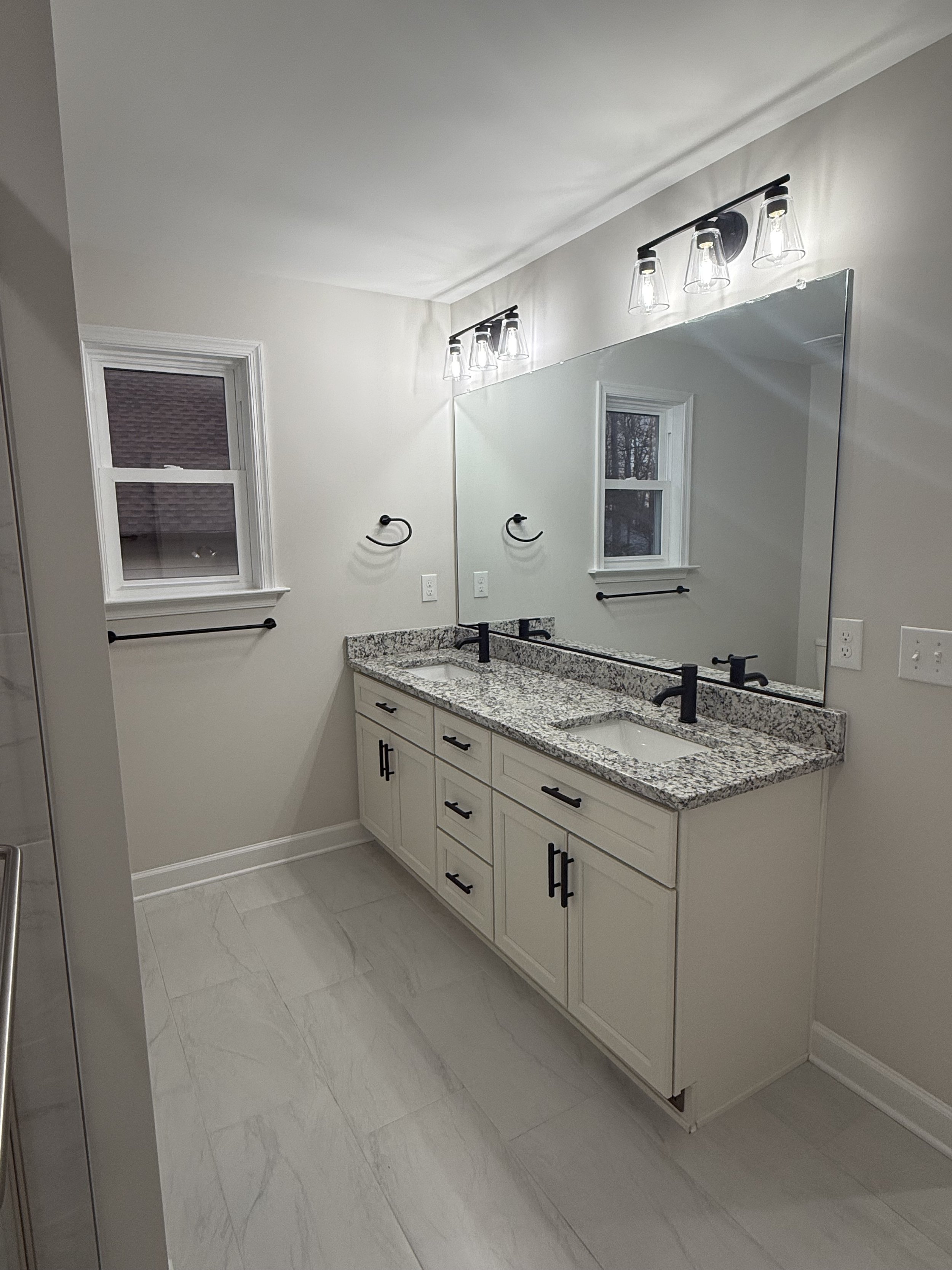 Modern bathroom with double vanity, granite countertop, black faucets, large mirror, and two small windows with white trim.