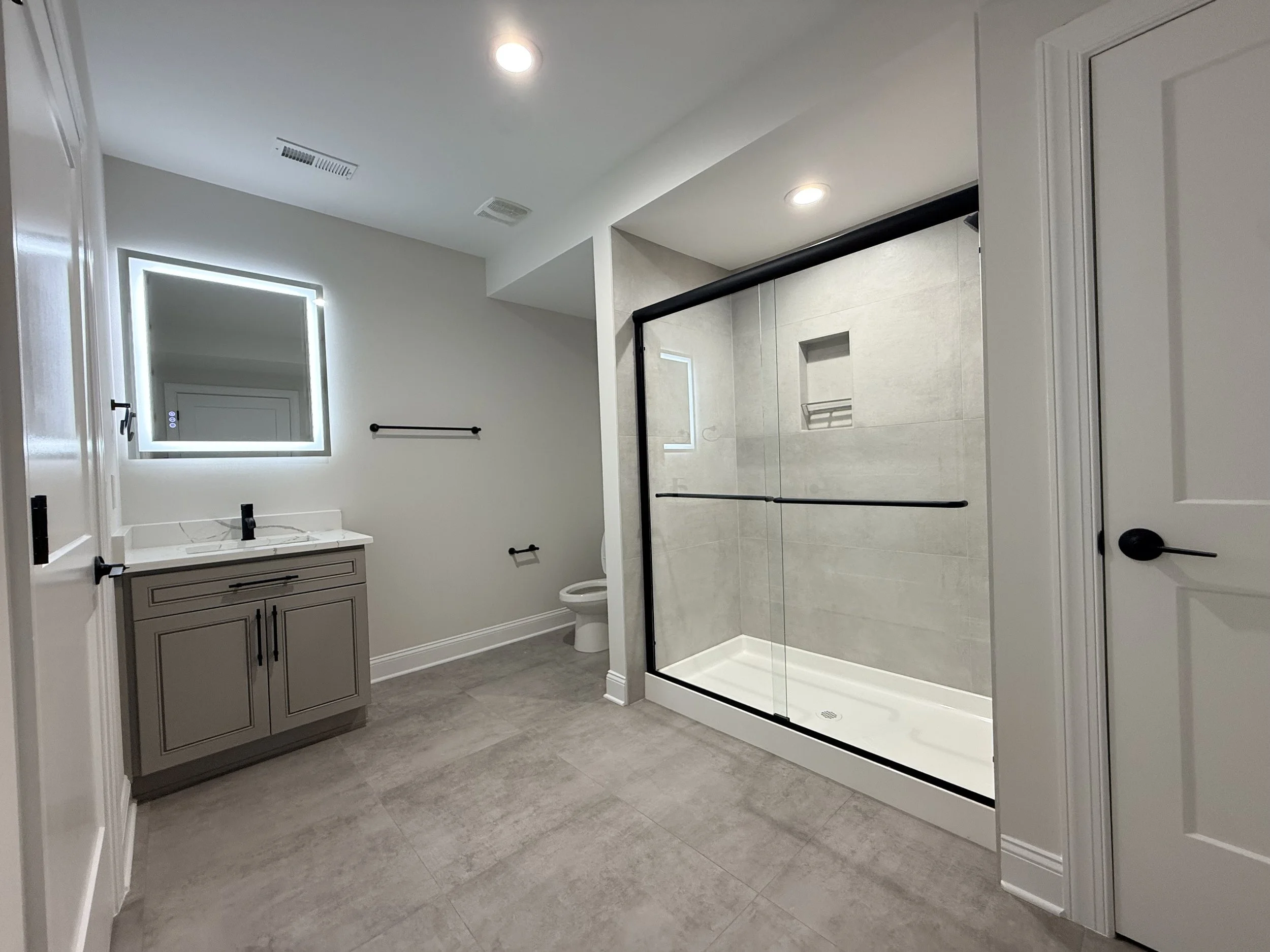 Modern bathroom with light grey floor tiles, a grey vanity with black hardware, a rectangular mirror with LED lighting, two small black towel bars, a toilet, and a large walk-in shower with a glass door, black accents, and built-in shelves.
