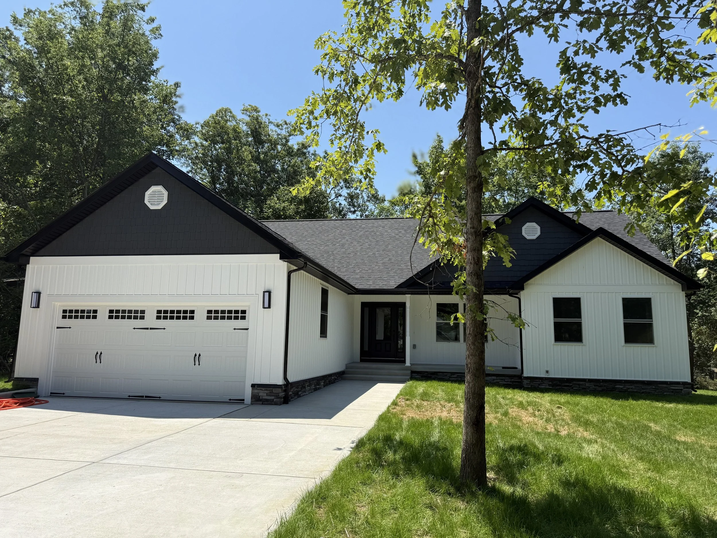 Front view of a modern house with a white exterior, black roof, and two-car garage, surrounded by green trees and a lawn under a clear blue sky.