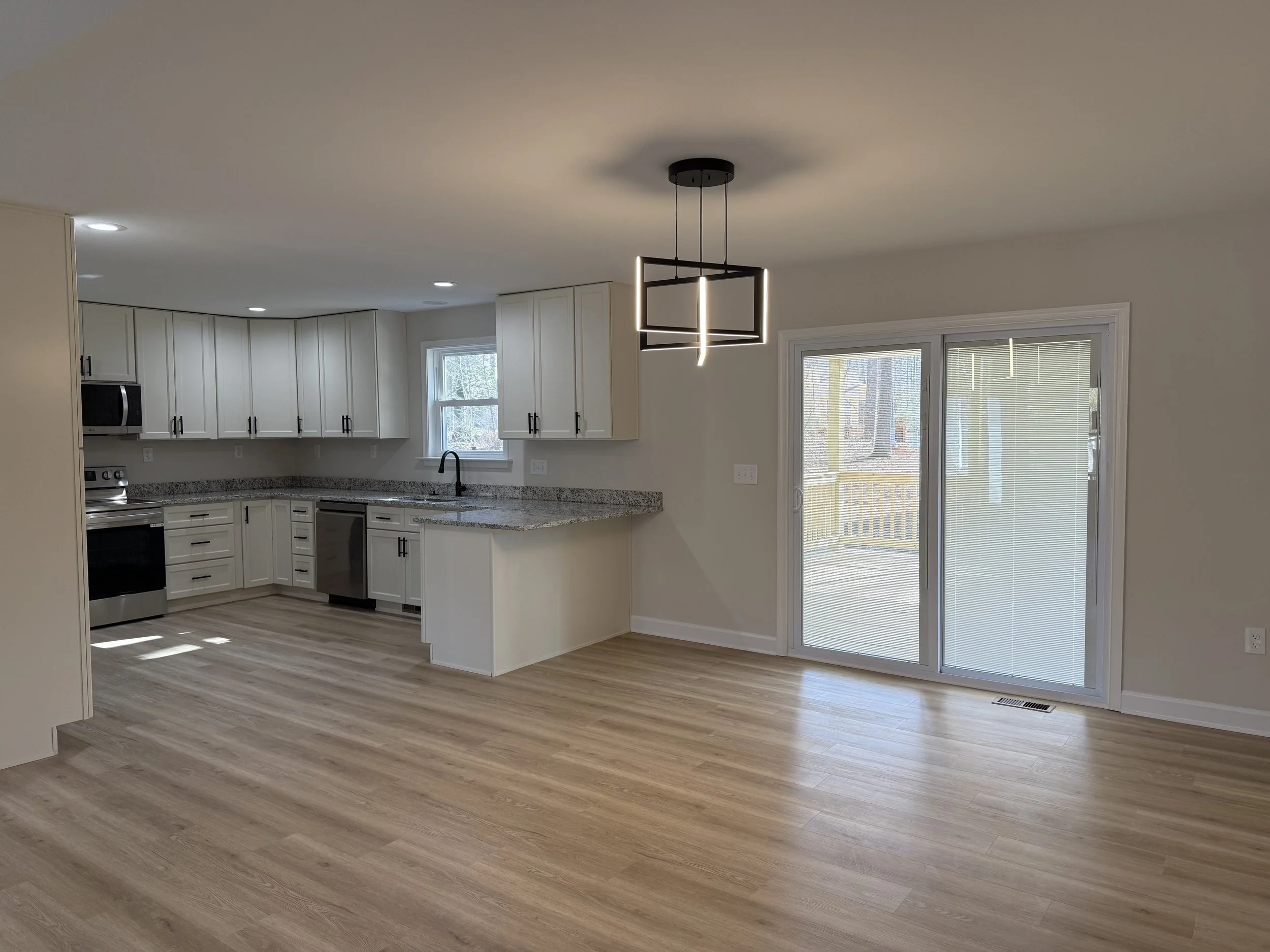 Empty kitchen and dining area with light hardwood floors, white cabinets, granite countertops, a sliding glass door with blinds, and modern black pendant lighting.