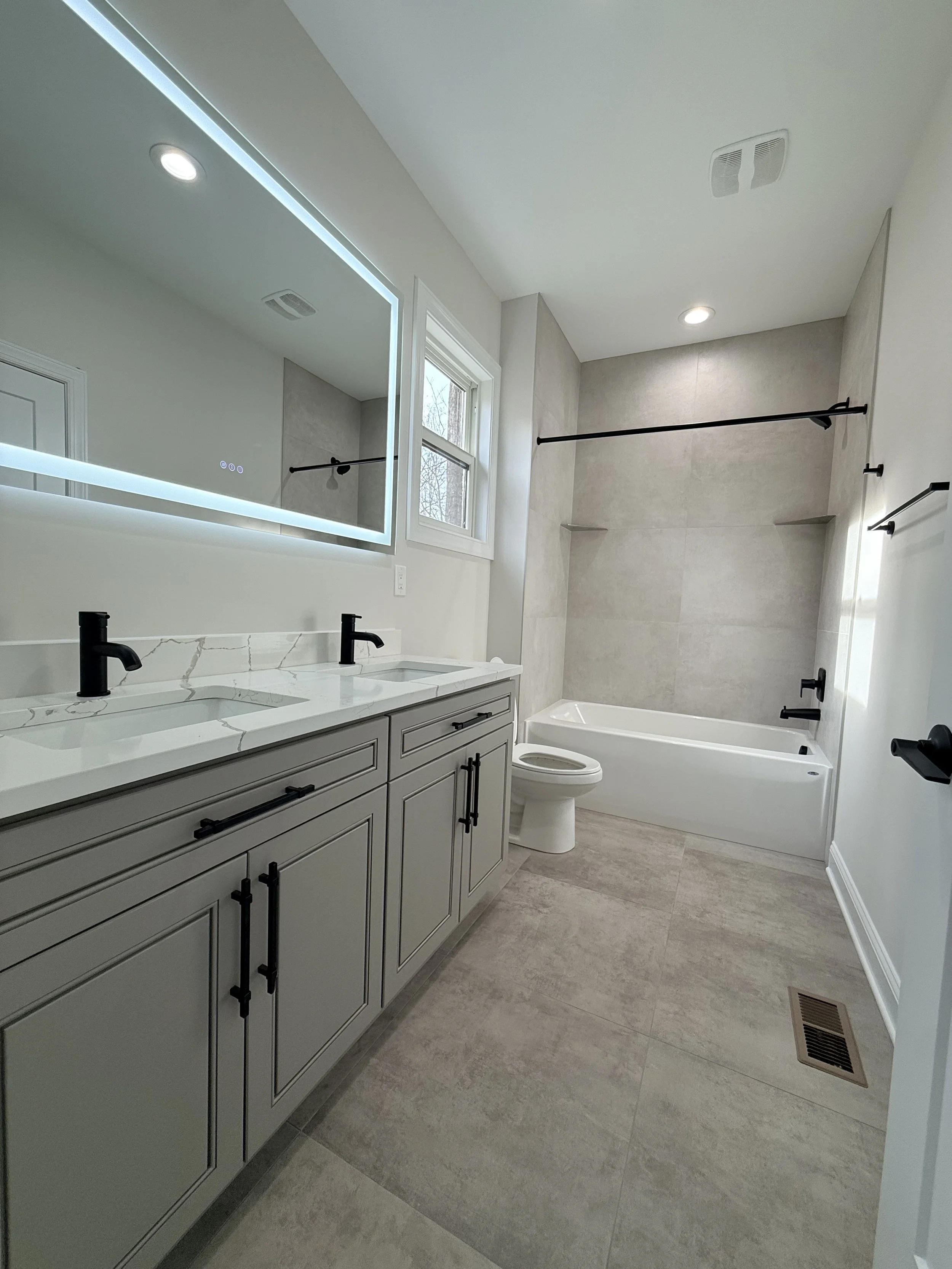 Modern bathroom with double vanity sinks, large mirror with backlighting, bathtub with shower area, toilet, beige floor tiles, gray and white walls, black fixtures, window, and ceiling lights.