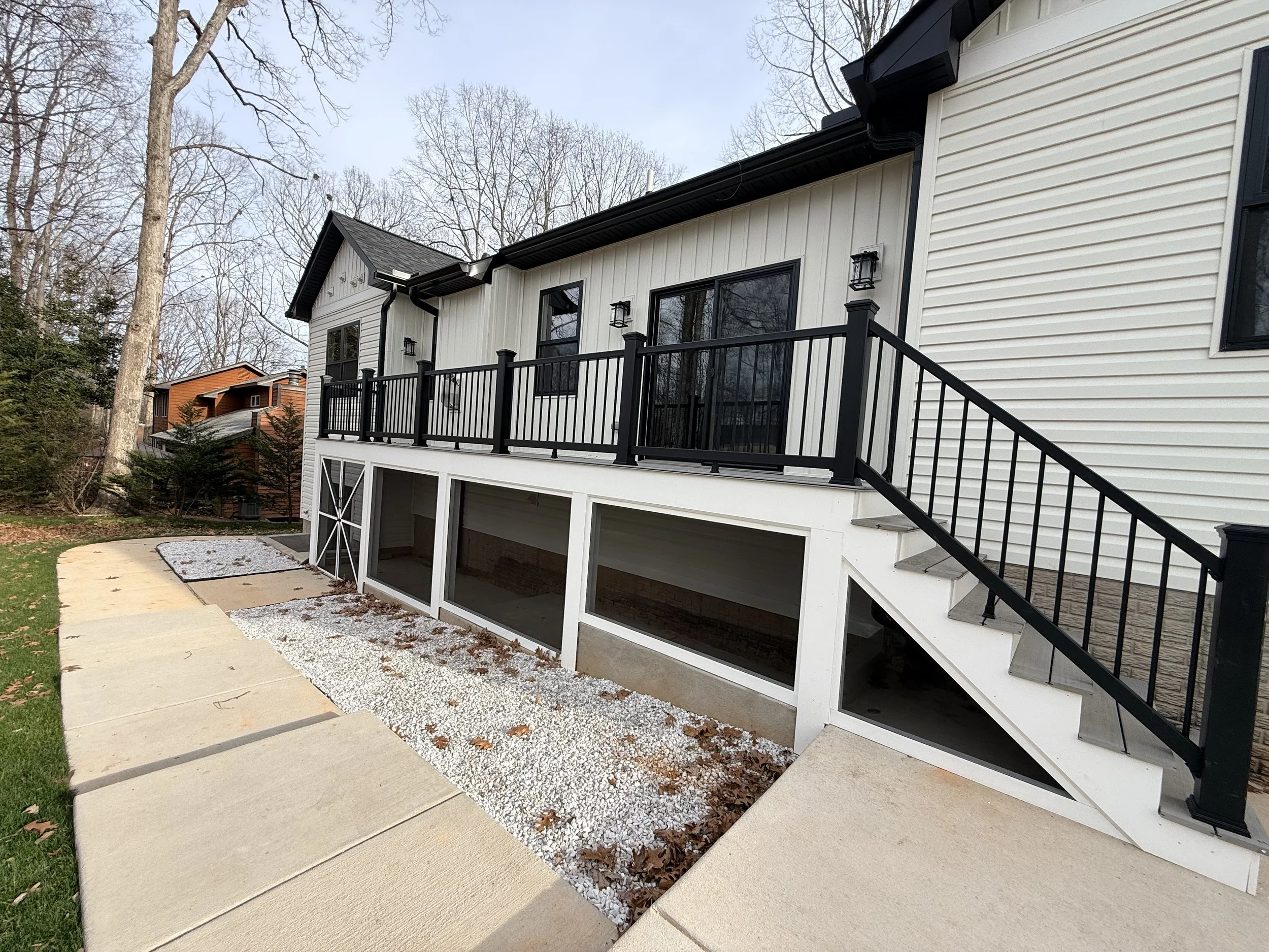 Back view of a modern white house with a black railing deck and stairs, with an open basement area underneath, surrounded by trees.