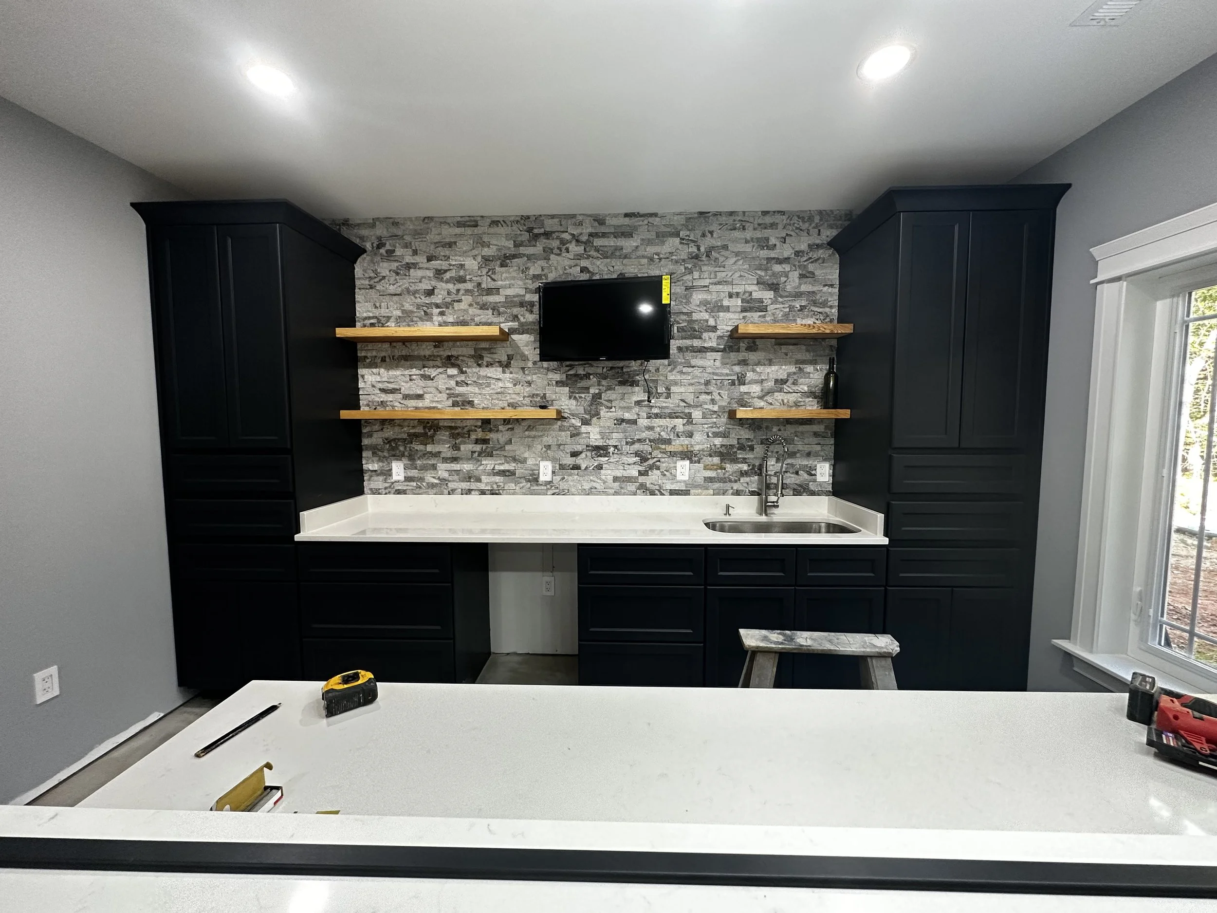 Kitchen with black cabinets, gray stone backsplash, white countertop, small TV mounted on the wall, open wooden shelves, sink, and window with trees outside.