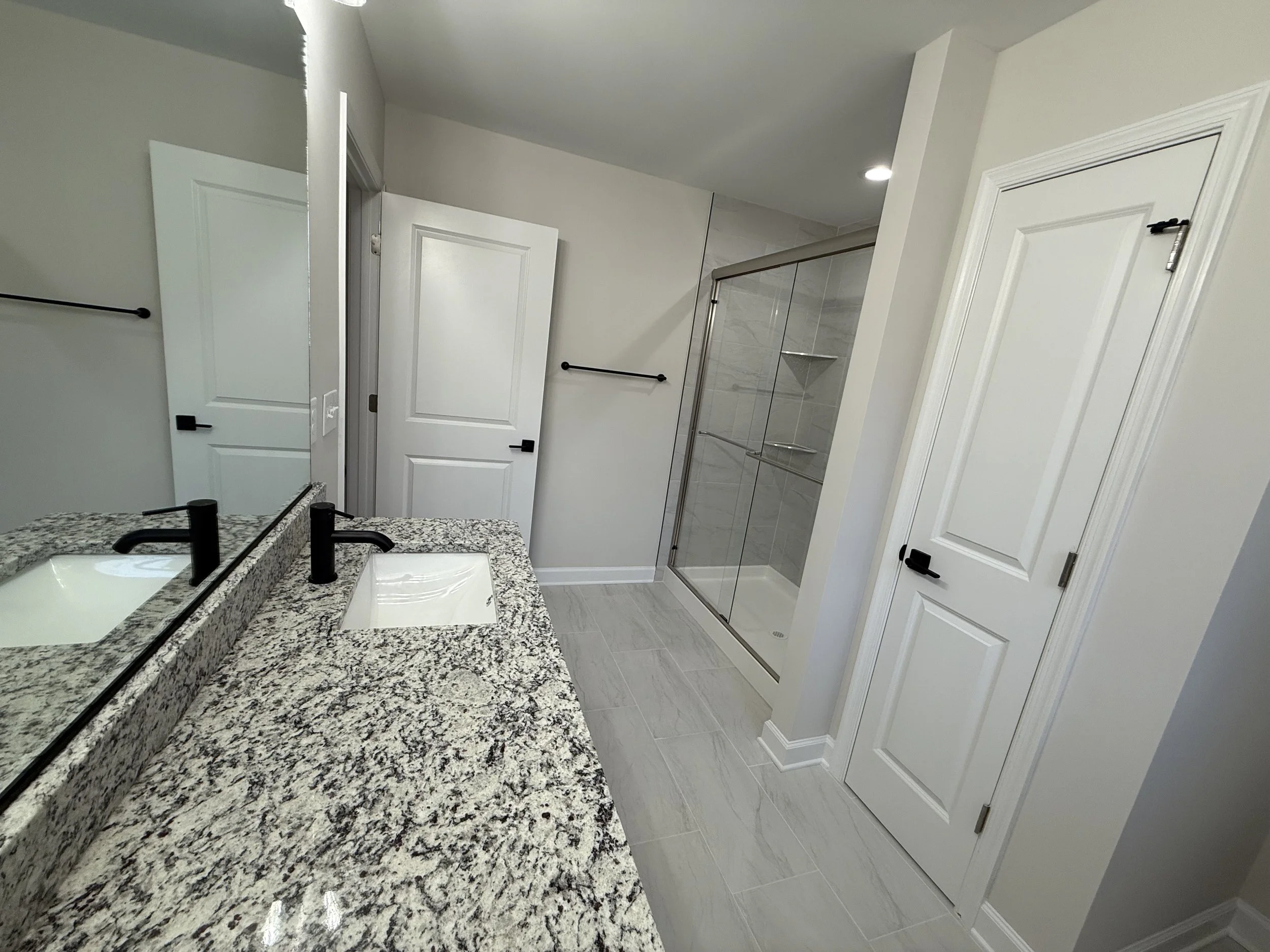 Modern bathroom with granite countertop double sink vanity, white cabinets, a glass-enclosed shower, and light-colored floor tiles.