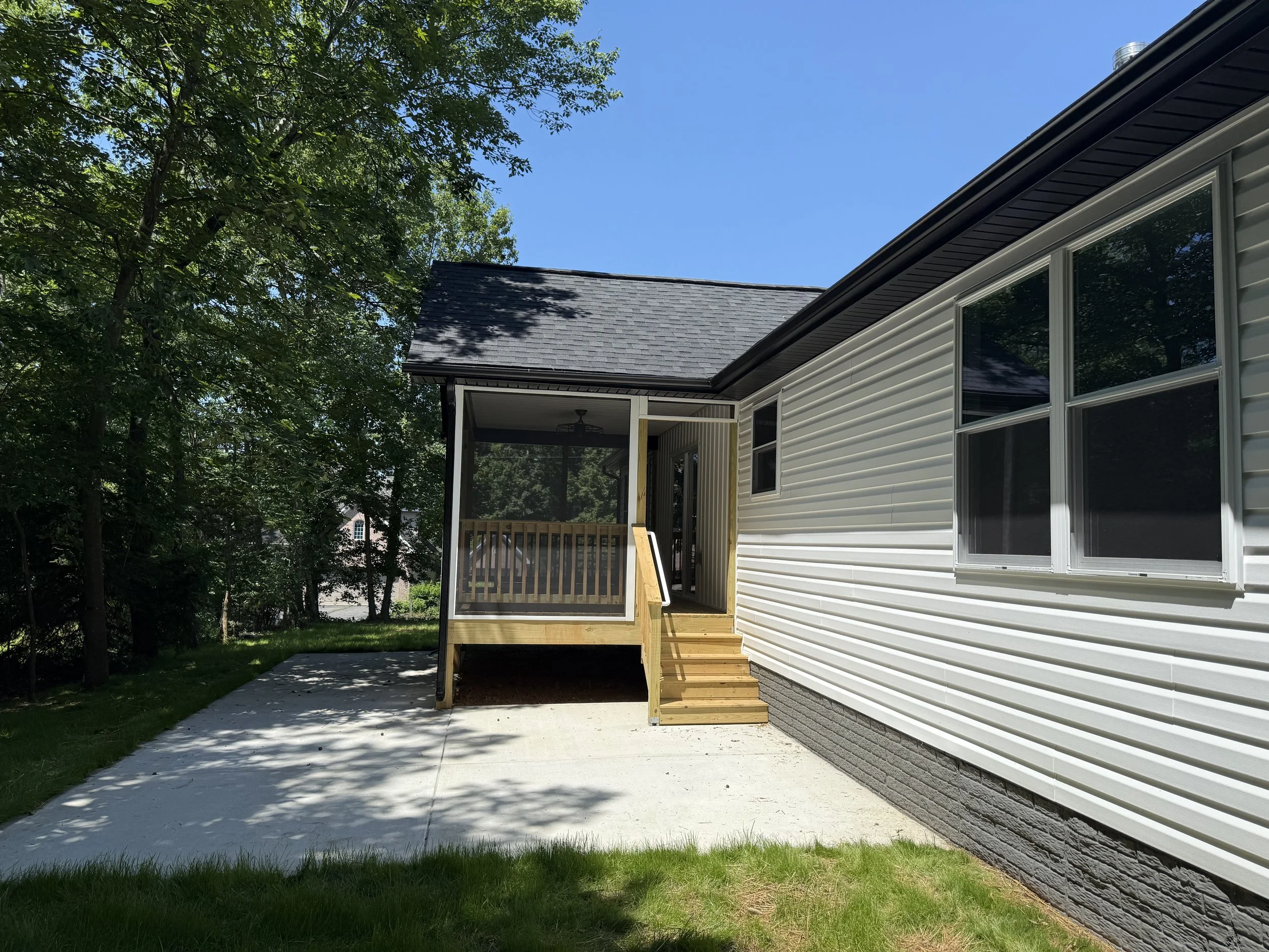 Backyard patio with a small wooden staircase leading to a screened porch attached to a white house, surrounded by trees.