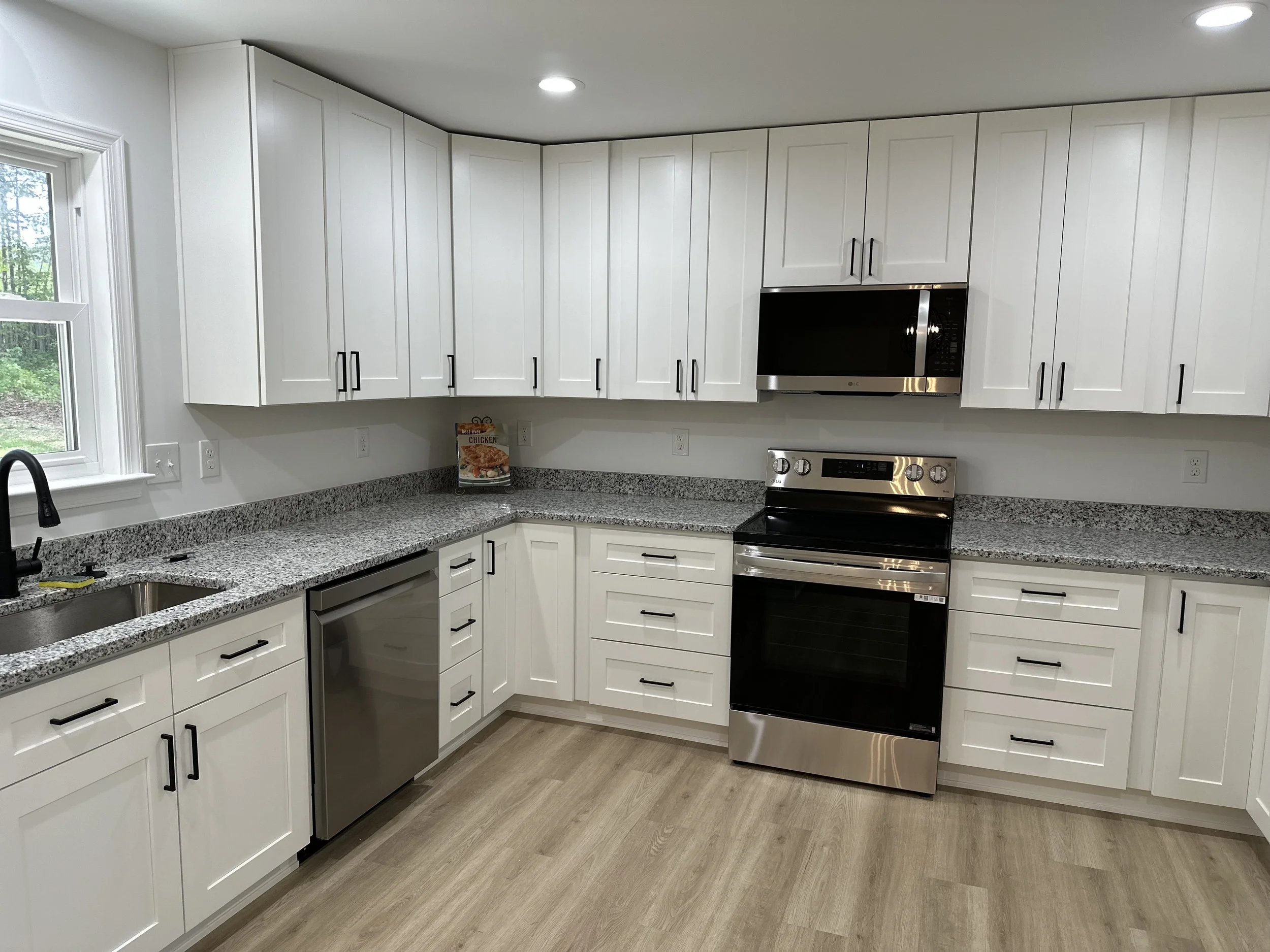 Modern kitchen with white cabinets, granite countertops, stainless steel appliances including a microwave, oven, and dishwasher, black faucet, and light wood flooring.