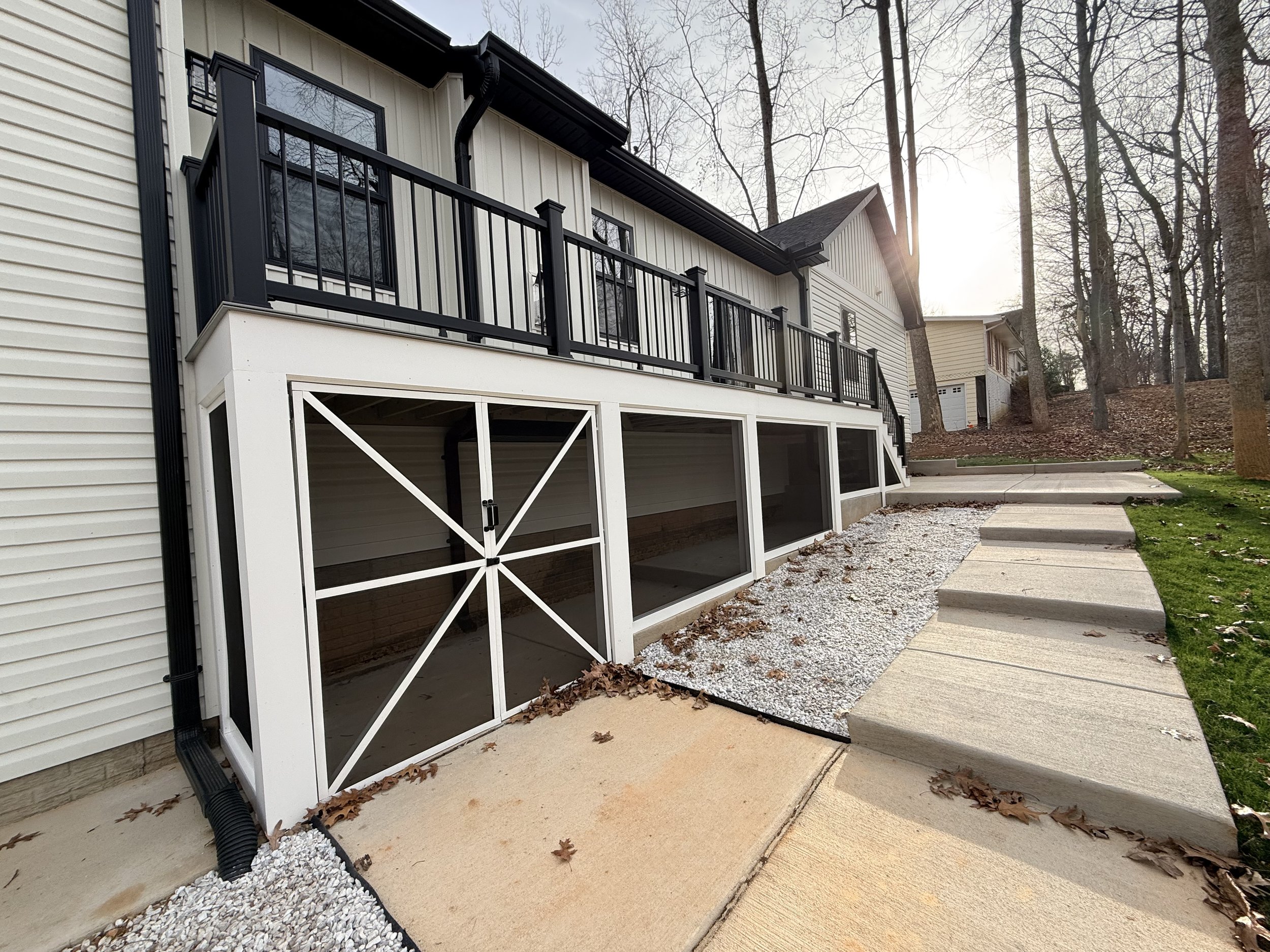 Backyard patio with a screened porch, concrete steps, gravel, and grass, with trees in the background and sunlight shining through.