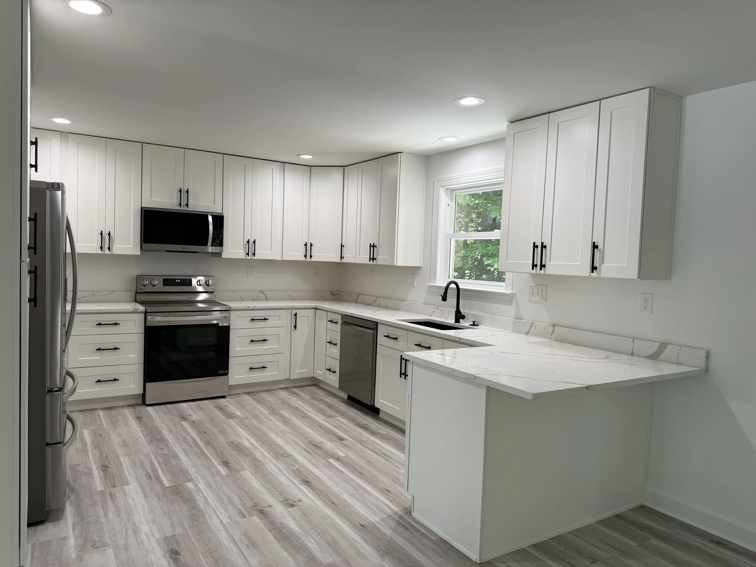 Modern kitchen with white cabinets, gray and white marble countertops, stainless steel appliances, a window with greenery outside, and light wood flooring.