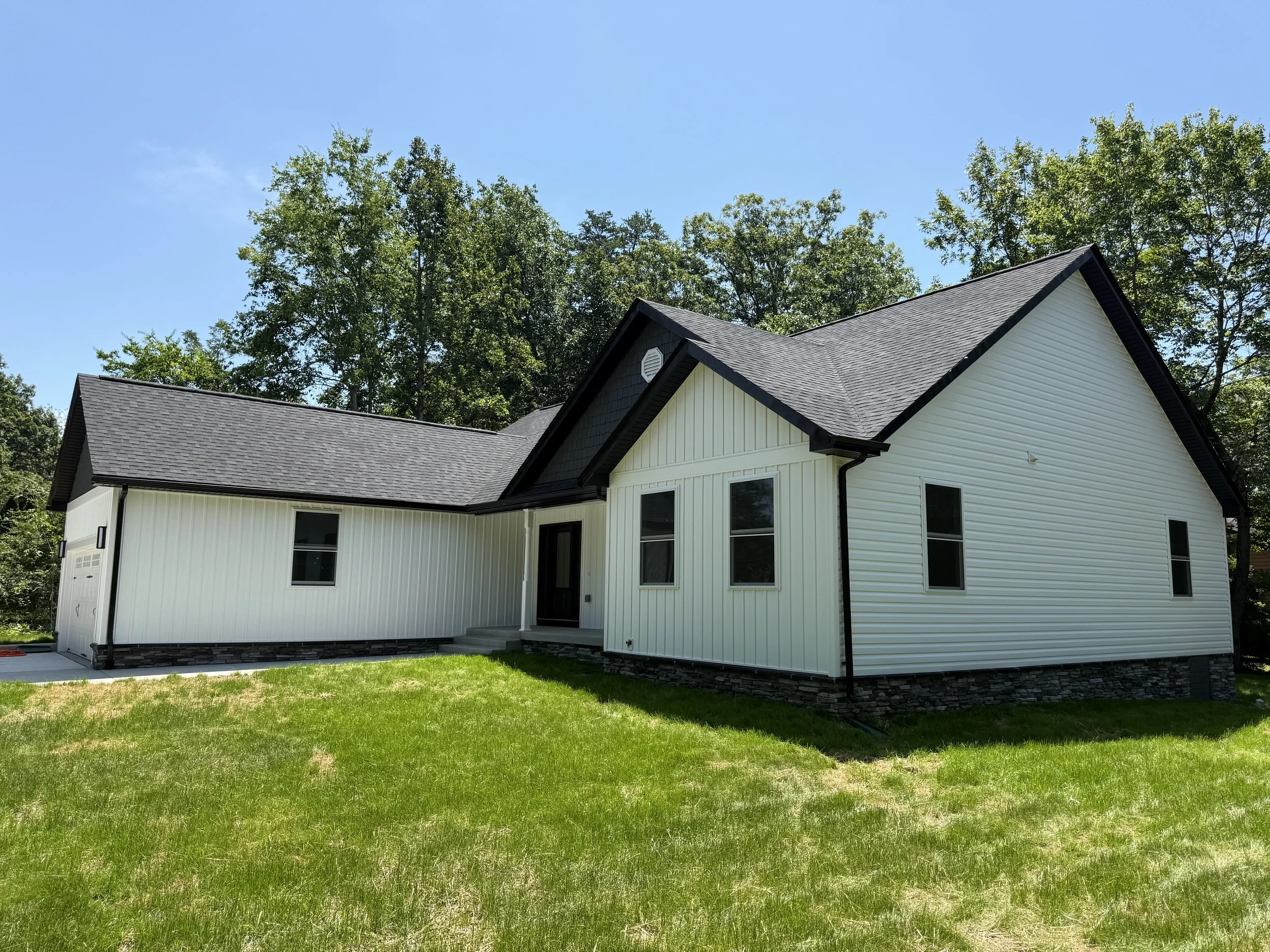 Modern white house with black roof and trim, situated on a grassy lawn, surrounded by trees under a clear blue sky.