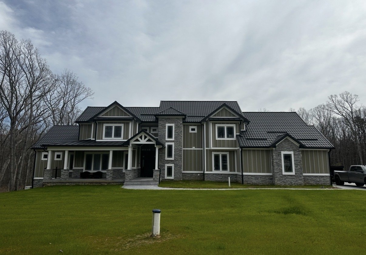 A large modern house with gray siding and stone accents, front porch with columns, well-maintained green lawn, and a driveway with a parked truck in the background.