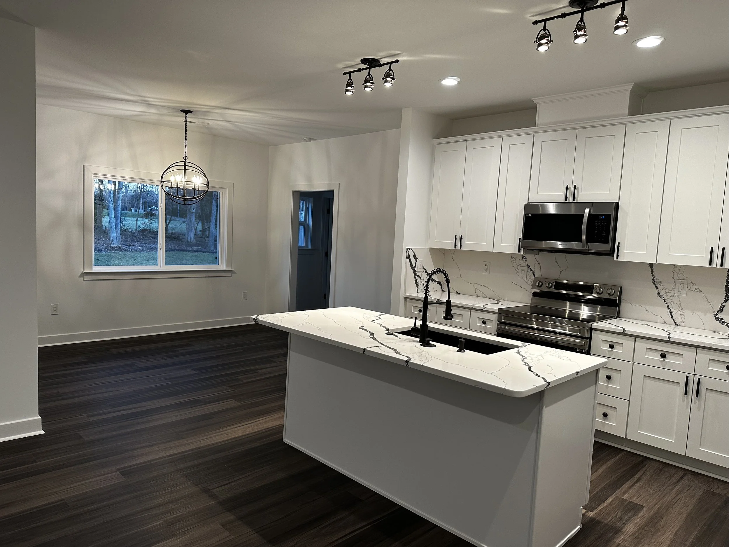 Modern kitchen with white cabinets, marble countertops, and stainless steel appliances, including a microwave and oven. An island with a marble top and black sink faucet is centered in the space. There is a large window providing natural light, and a