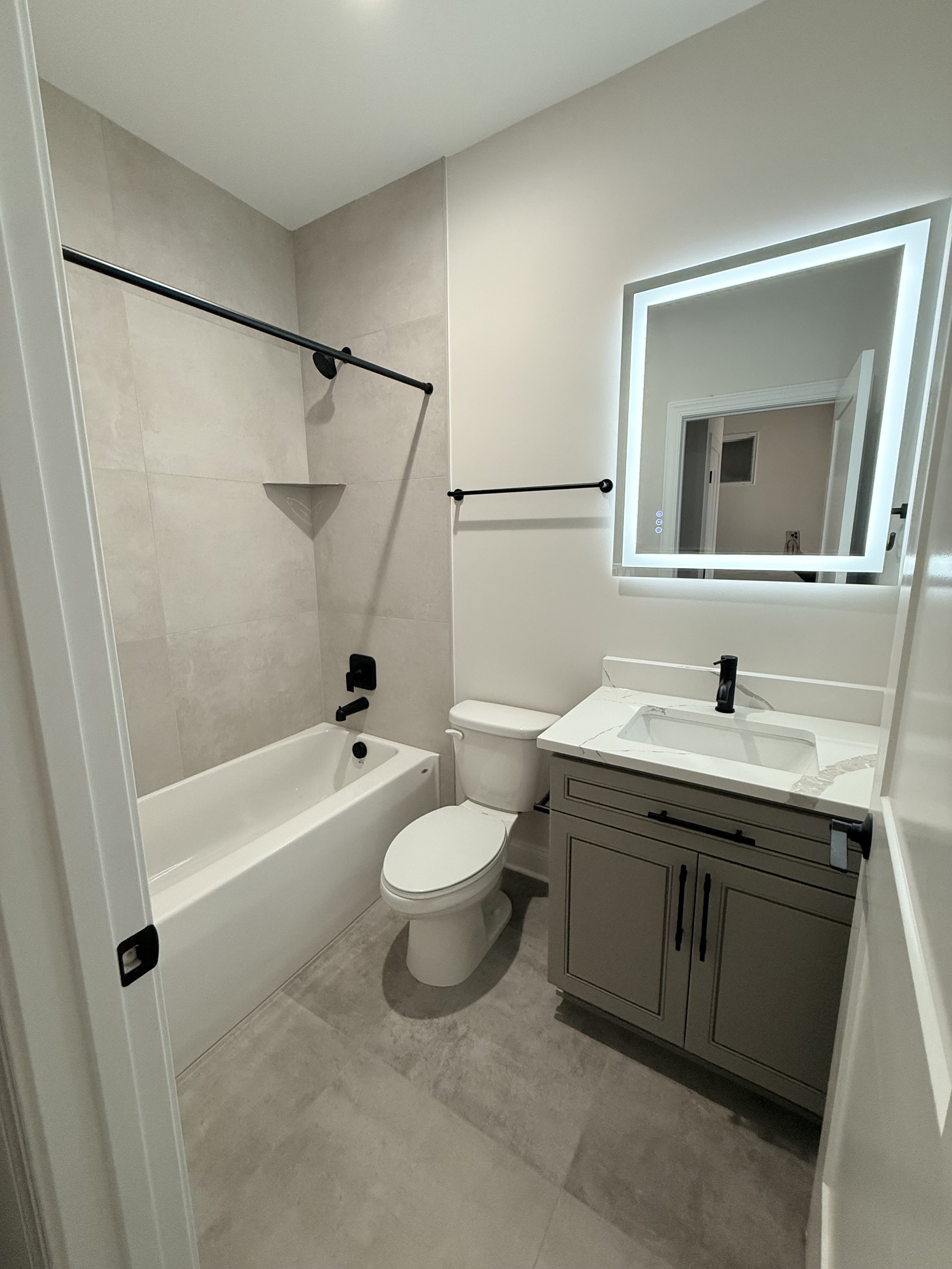 A modern bathroom featuring a bathtub with a black shower rod, a toilet, a vanity with a white marble countertop and a rectangular sink, and a large illuminated square mirror.