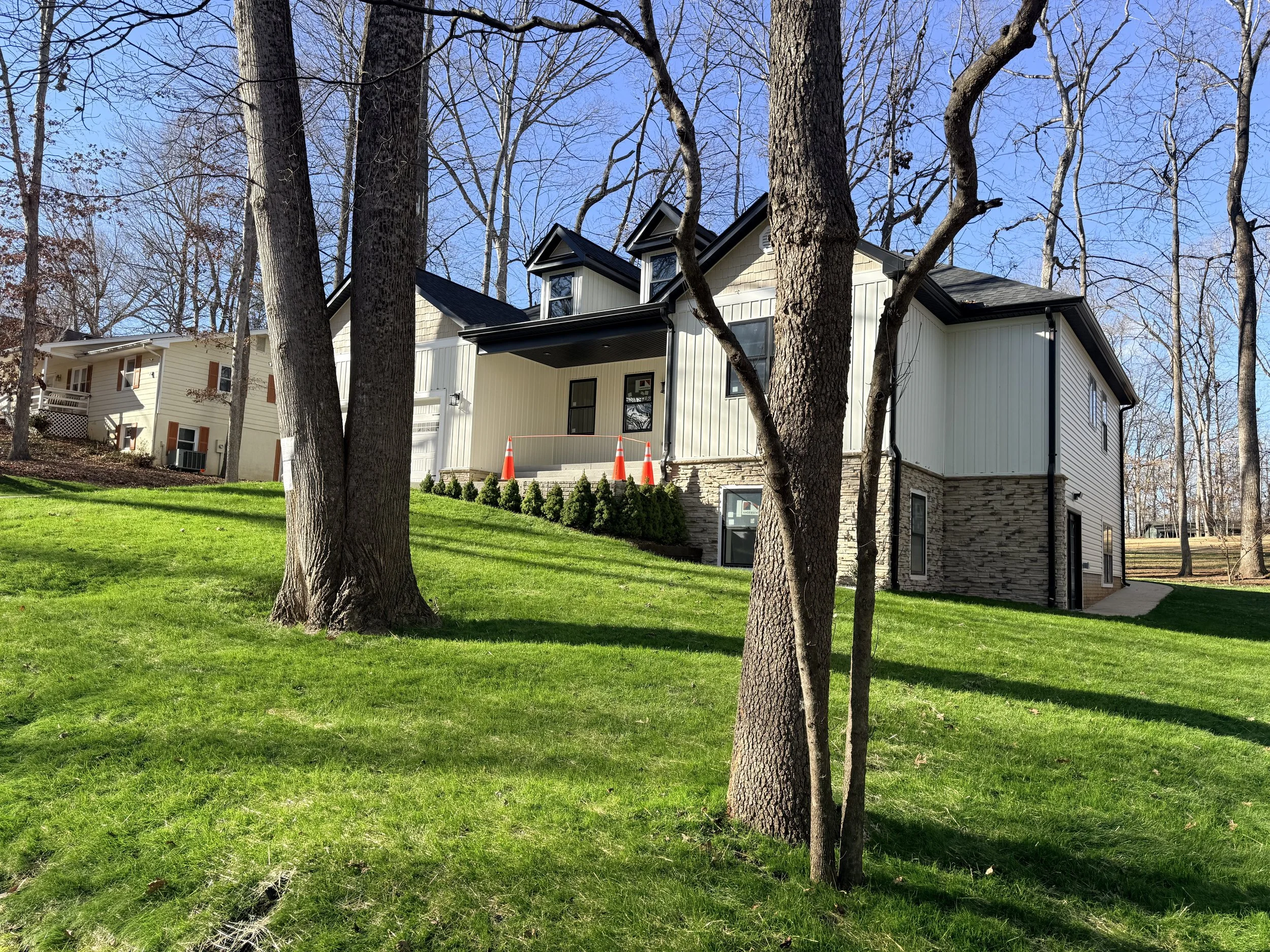 A modern house with cream-colored siding and stone accents, situated on a grassy hill surrounded by tall trees, with caution cones placed near the front porch.
