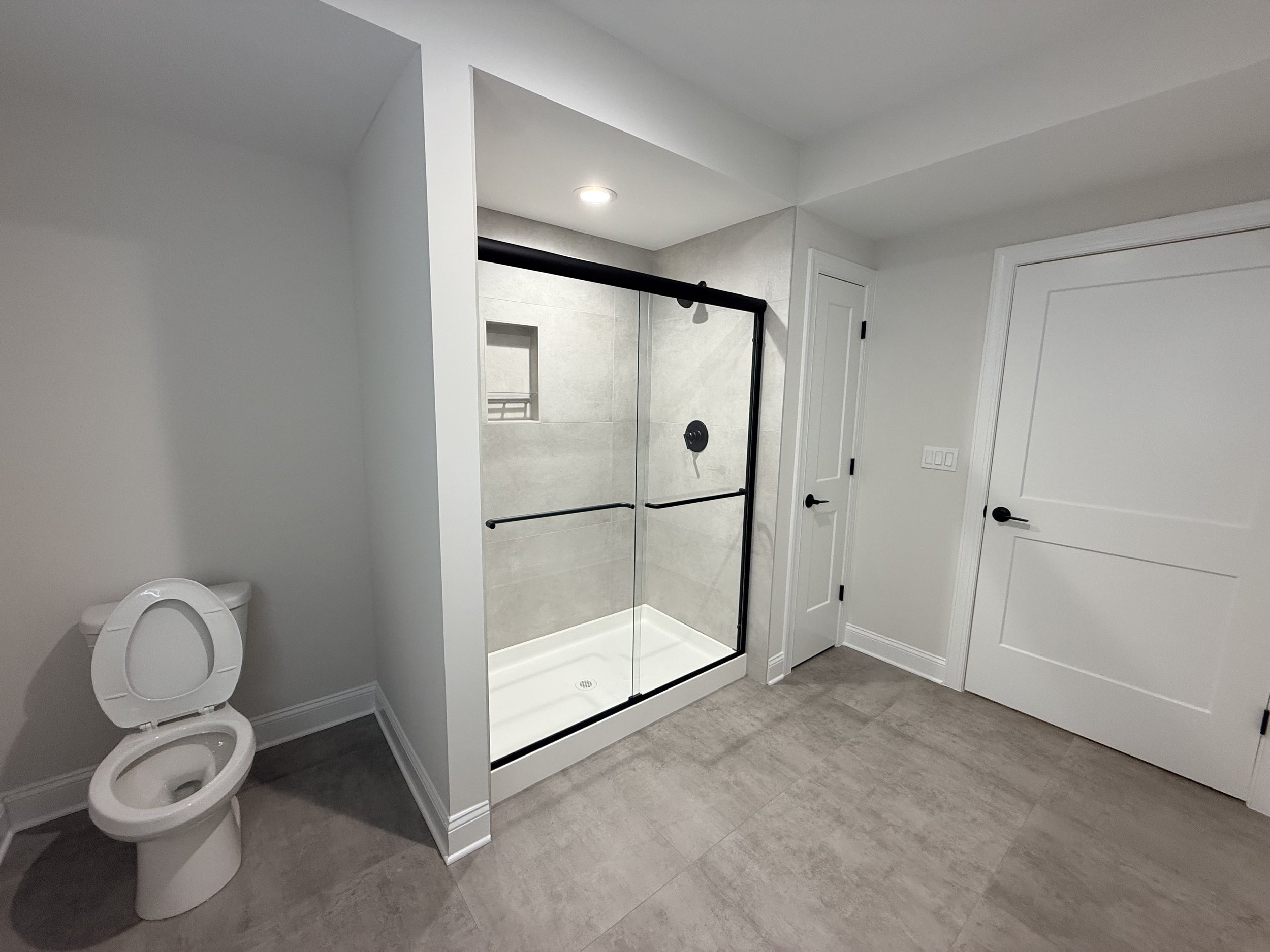 A bathroom with a white toilet on the left, a glass shower with black hardware in the center, and two white doors, one of which is partially open, on the right. The bathroom has beige tiled flooring and white walls.
