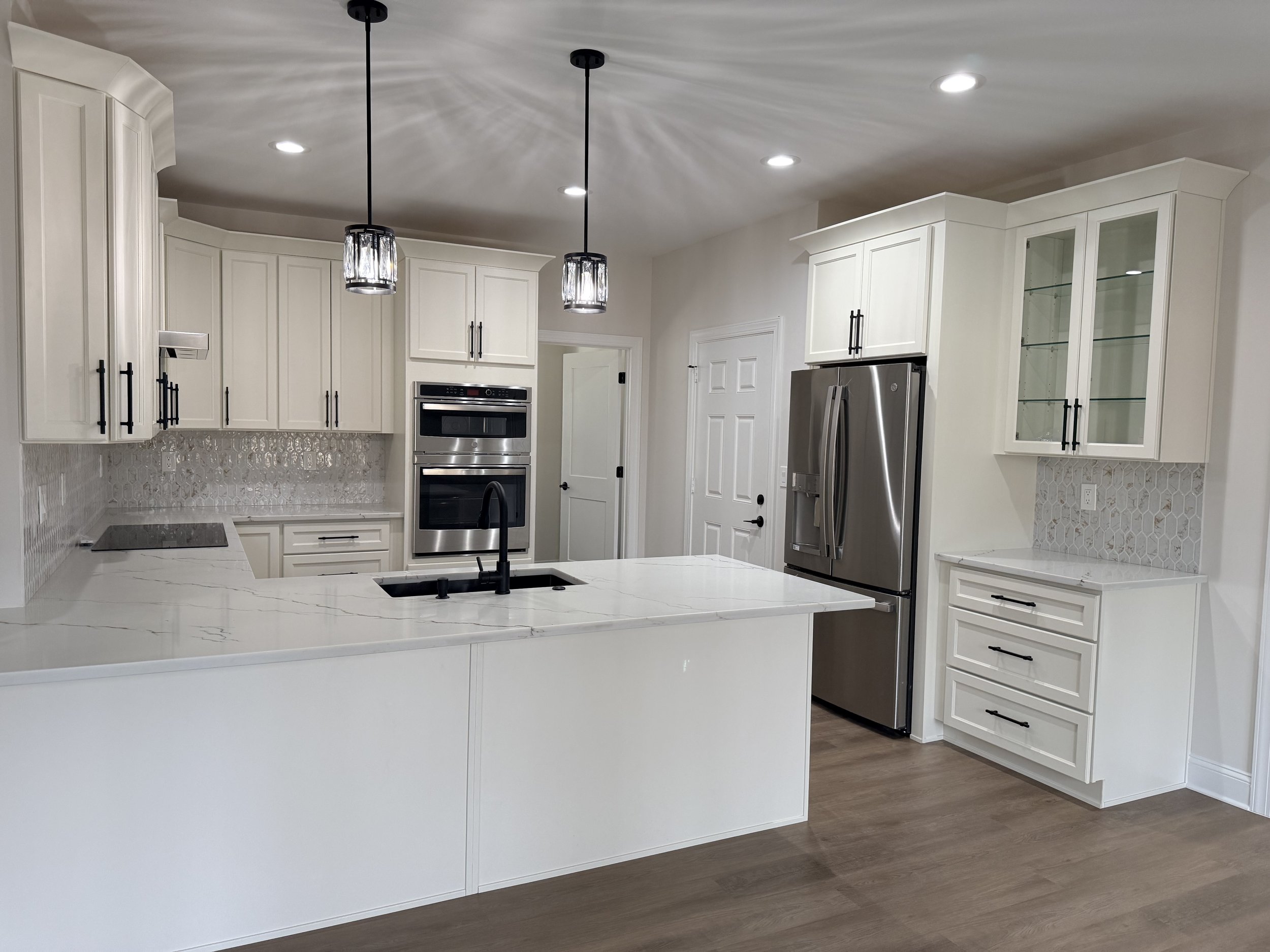 Modern kitchen with white cabinetry, marble countertops, stainless steel appliances including a refrigerator, double oven, and electric cooktop. Pendant lights hang over the island, and there is a decorative backsplash.