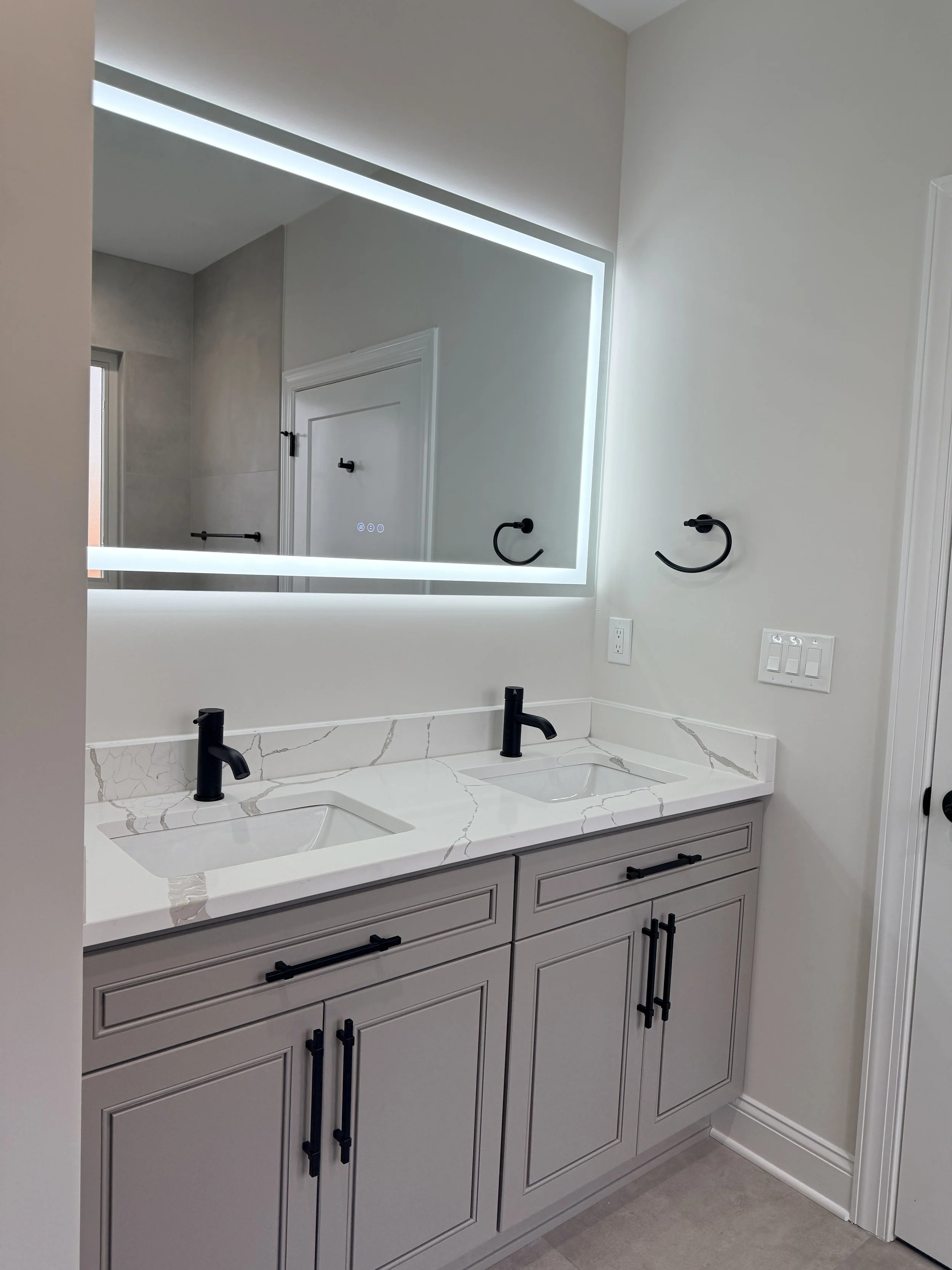 Modern bathroom vanity with dual sinks, black faucets, white marble countertop, and large backlit mirror.