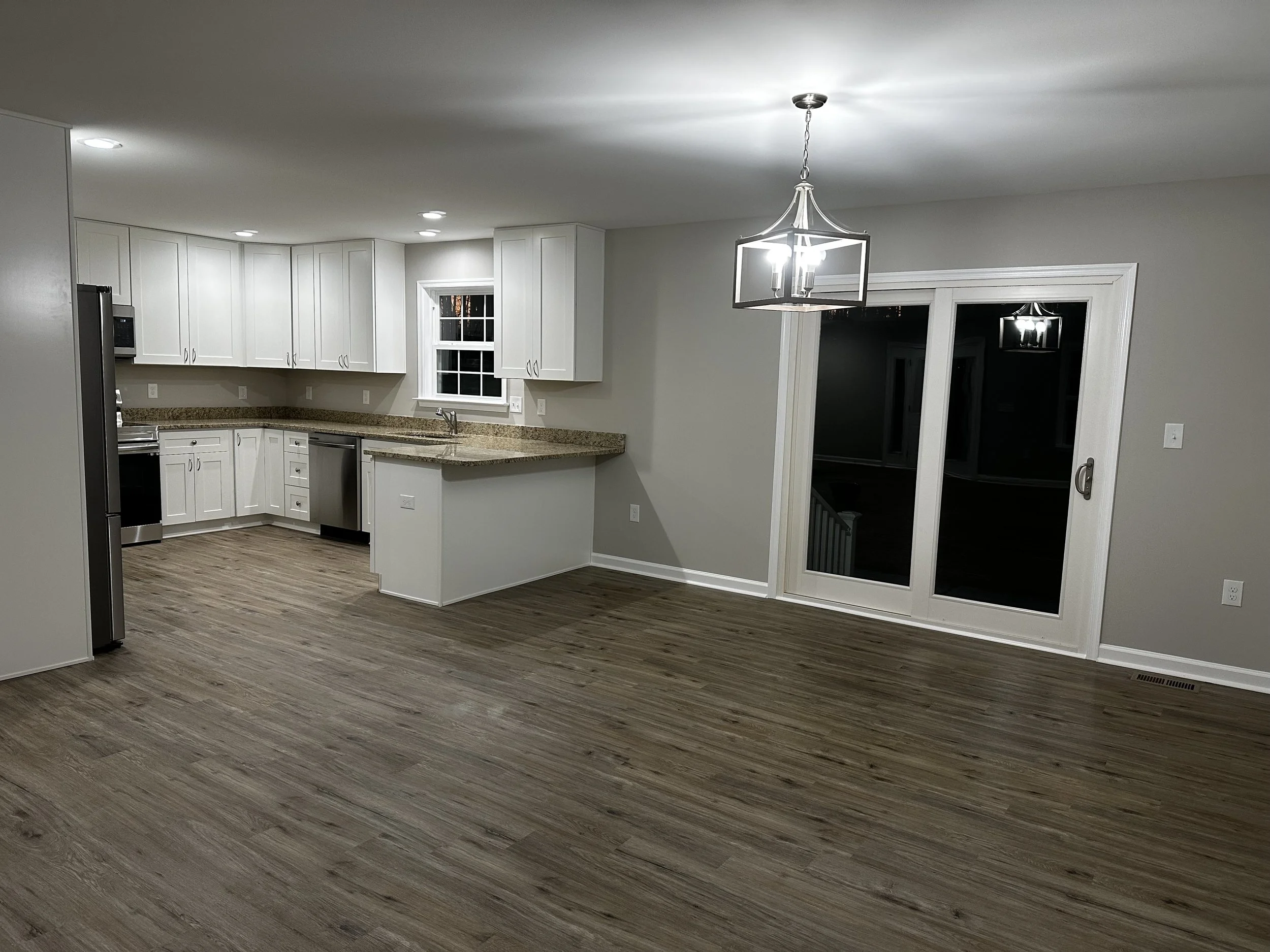 An empty kitchen and dining area with white cabinets, granite countertops, a window in the kitchen, and a sliding glass door leading outside. The room has hardwood floors and a modern light fixture hanging from the ceiling.