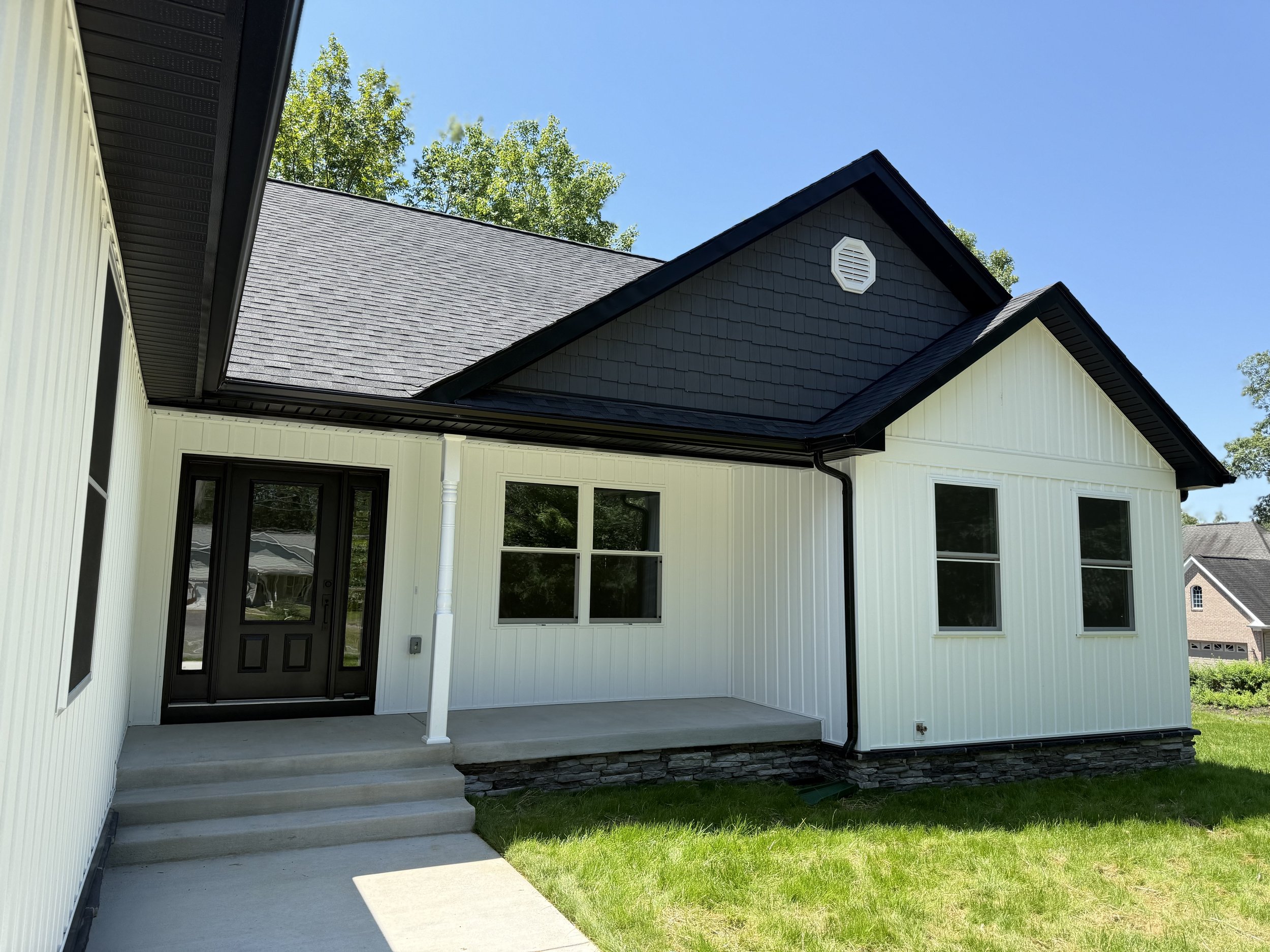 Front view of modern white house with black door and trim, gray roof, small porch with steps, on a sunny day with green grass and trees in the background.