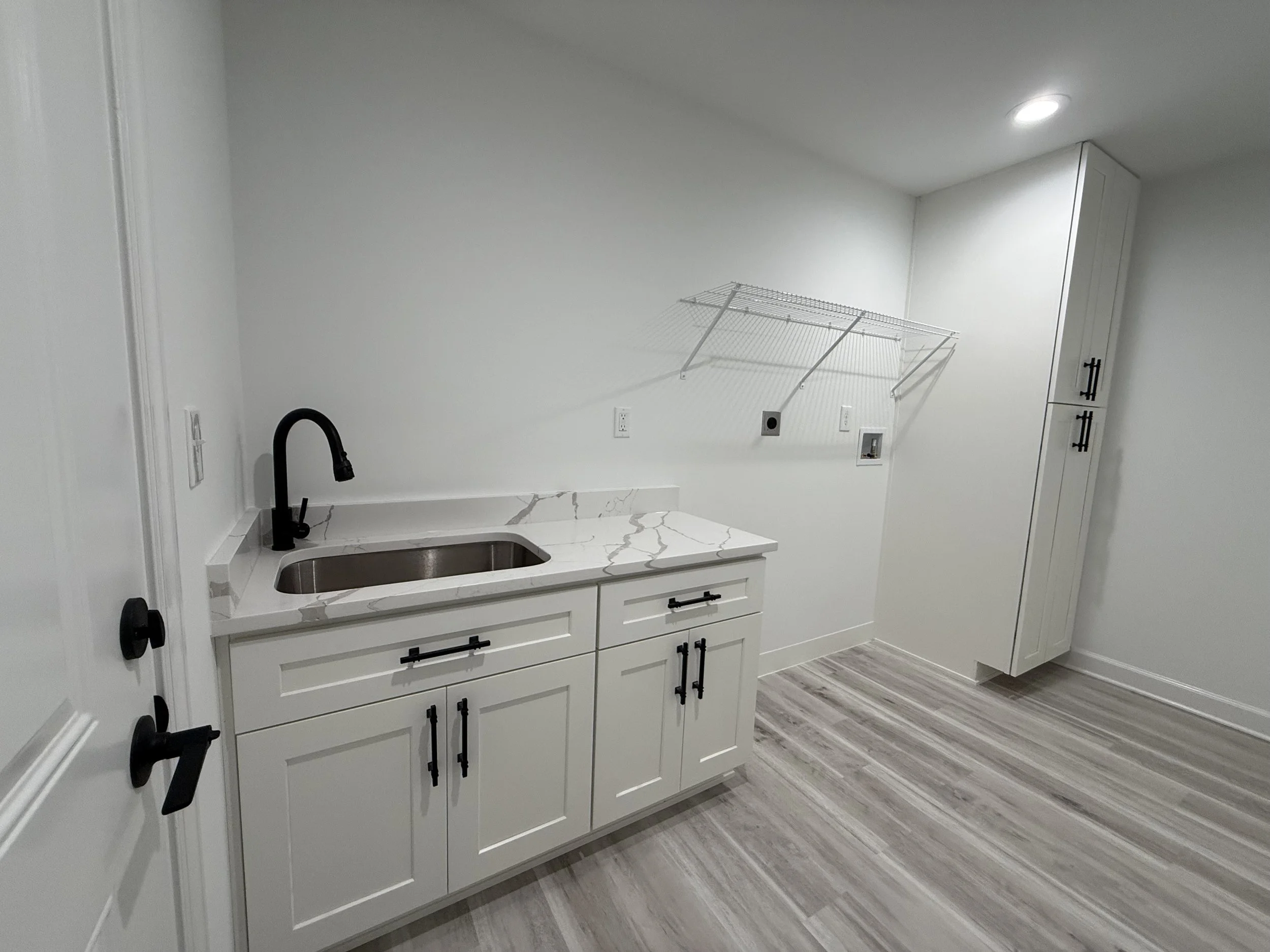 Empty laundry room with white cabinets, black faucet, marble countertop, wire shelf, and wood-look flooring.