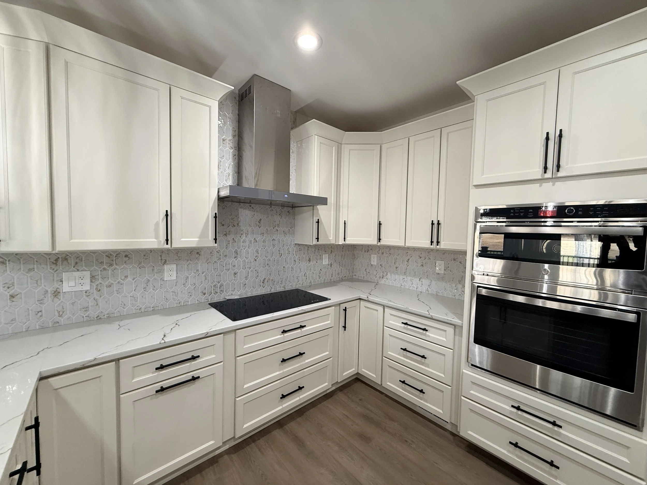 Modern kitchen with white cabinetry, black handles, stainless steel double oven, black cooktop, textured backsplash, and wood flooring.